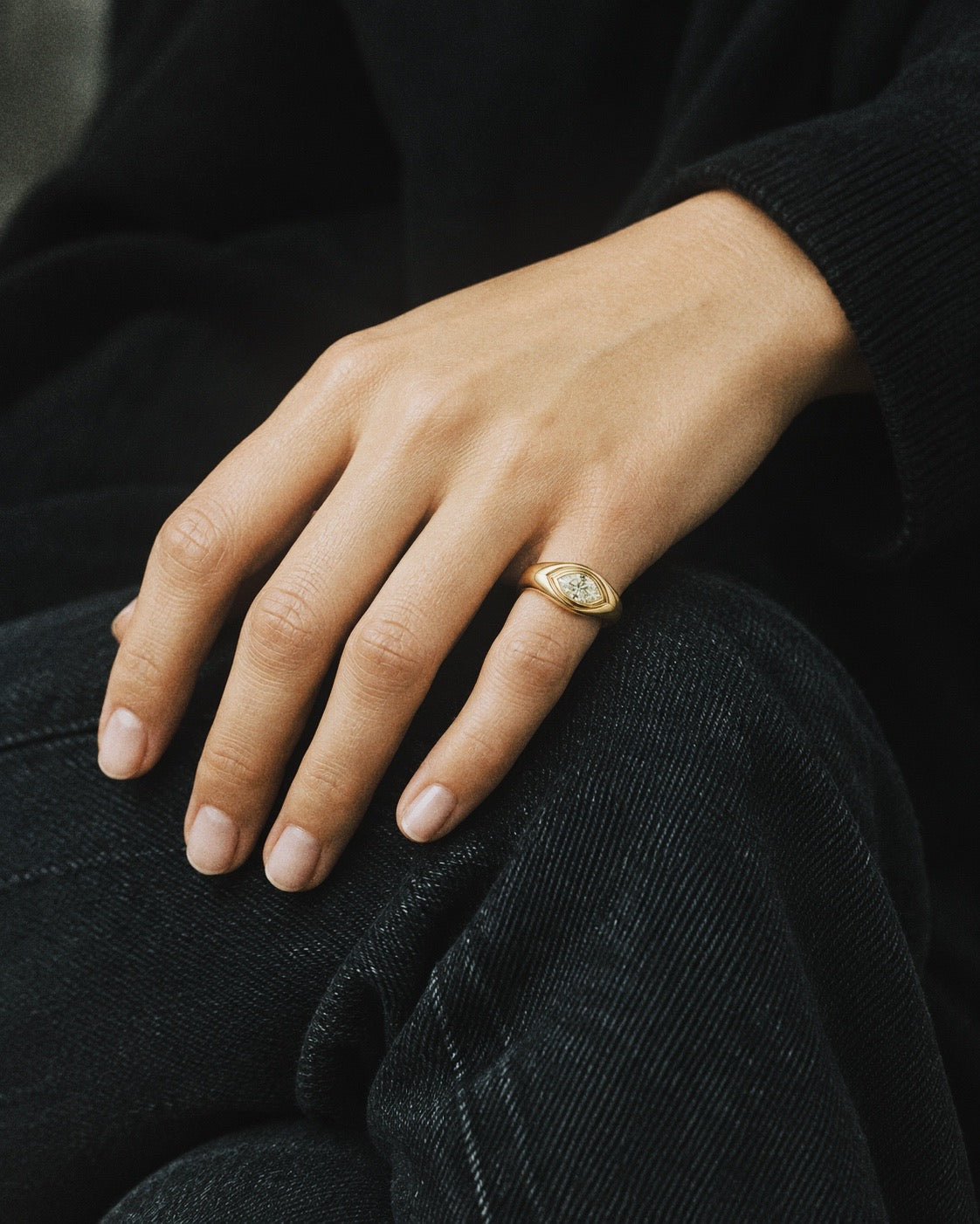 A close-up of a person's hand resting on dark denim, wearing a gold ring with a white oval stone.