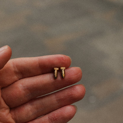 Two small gold earrings held in a hand against a blurred background