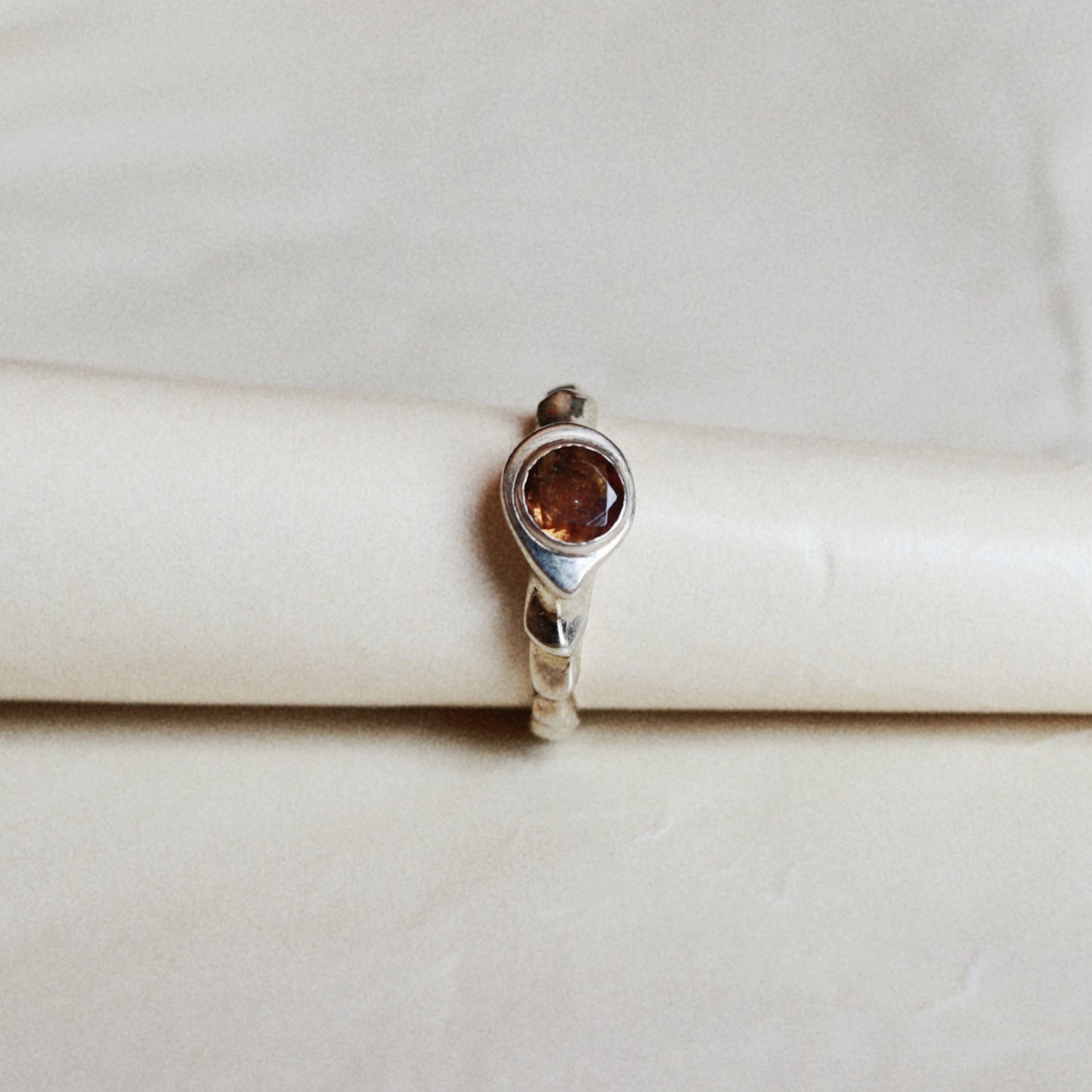 A silver ring with a round amber-brown gemstone, displayed on a cream-colored ring stand against a light beige background.