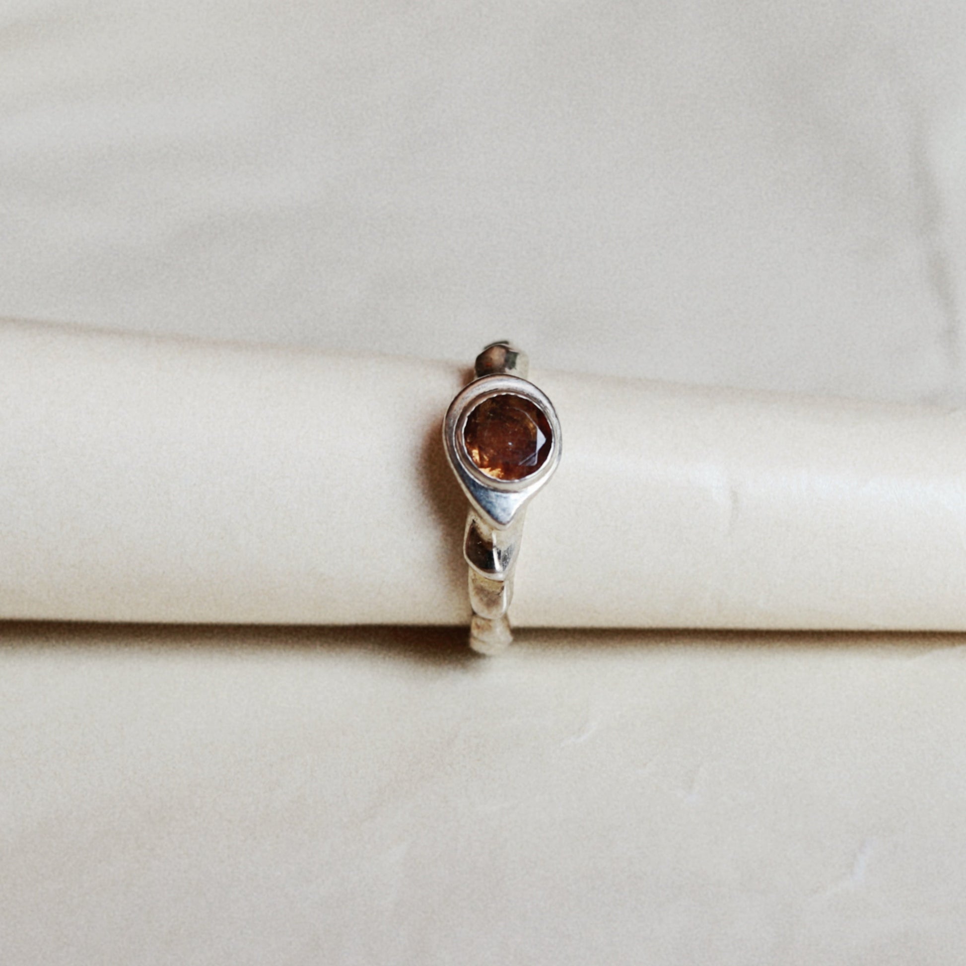 A silver ring with a round amber-brown gemstone, displayed on a cream-colored ring stand against a light beige background.