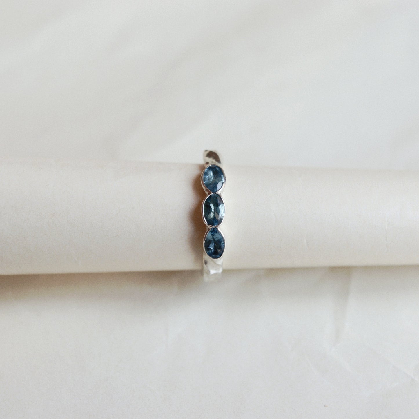 A silver ring featuring three oval-cut dark blue gemstones, displayed on a cream-colored cylindrical stand against a soft, light beige background.