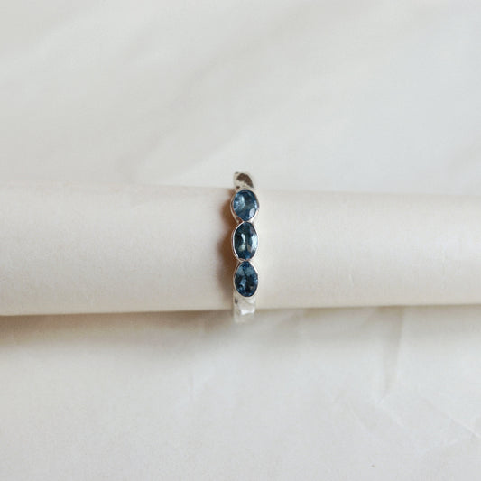 A silver ring featuring three oval-cut dark blue gemstones, displayed on a cream-colored cylindrical stand against a soft, light beige background.