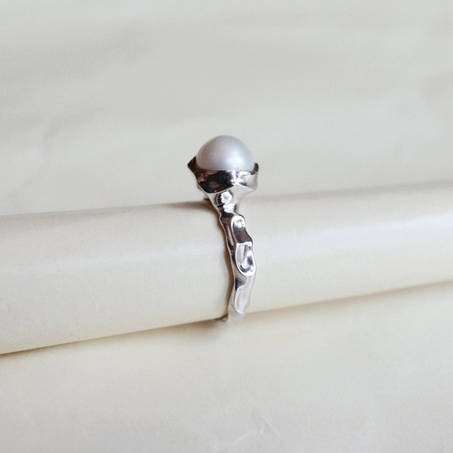 A silver ring with a round white pearl on a cream-colored display stand.