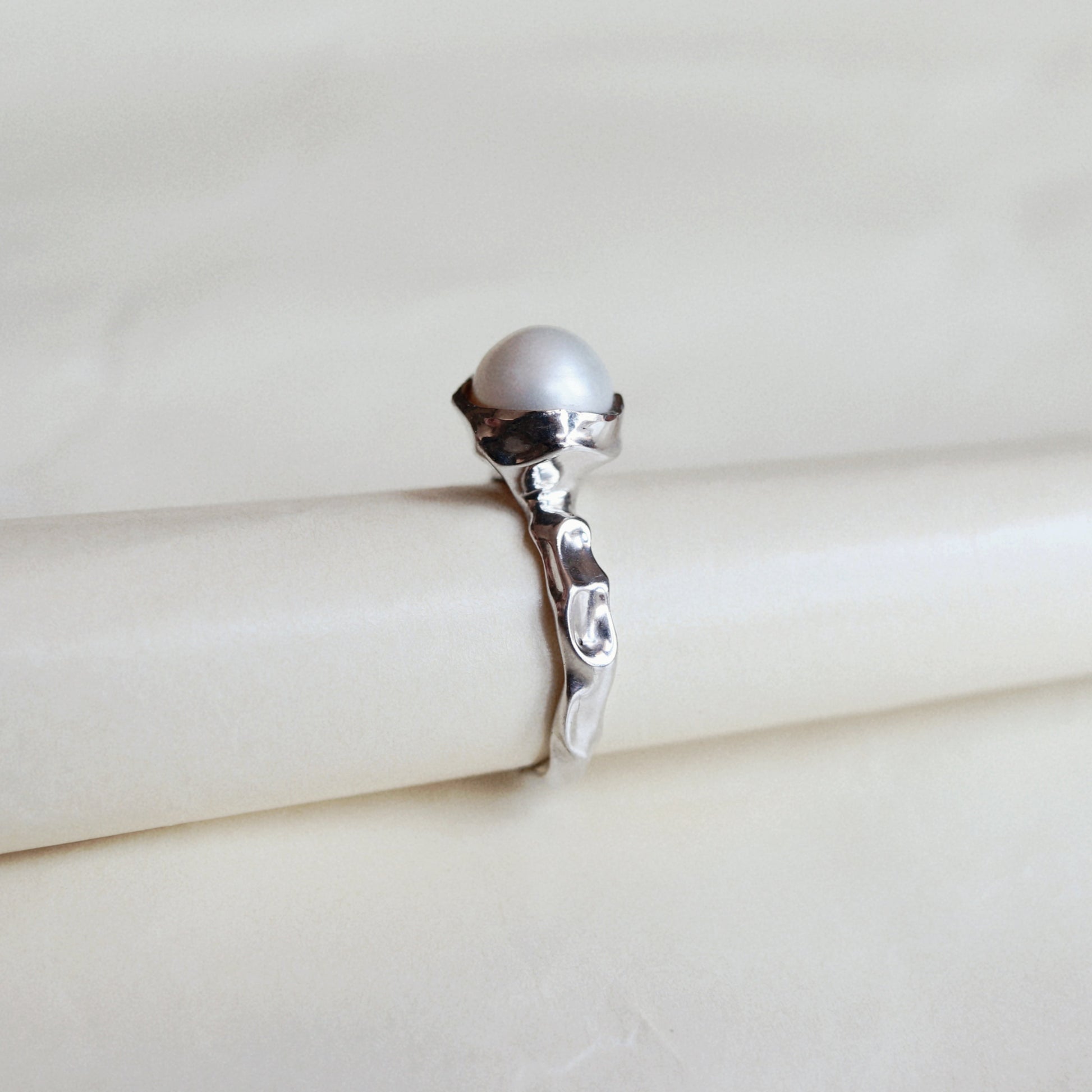 A silver ring with a round white pearl on a cream-colored display stand.