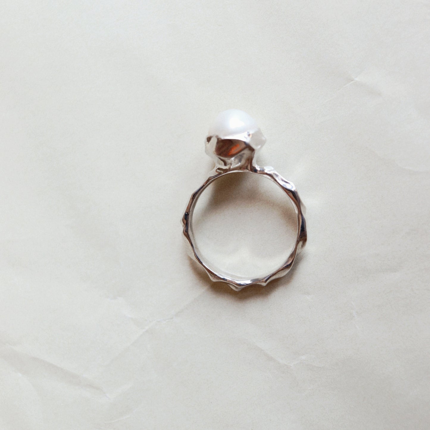 A silver ring with a textured, hammered band and a single white pearl set in a polished bezel, photographed on a wrinkled off-white paper background.