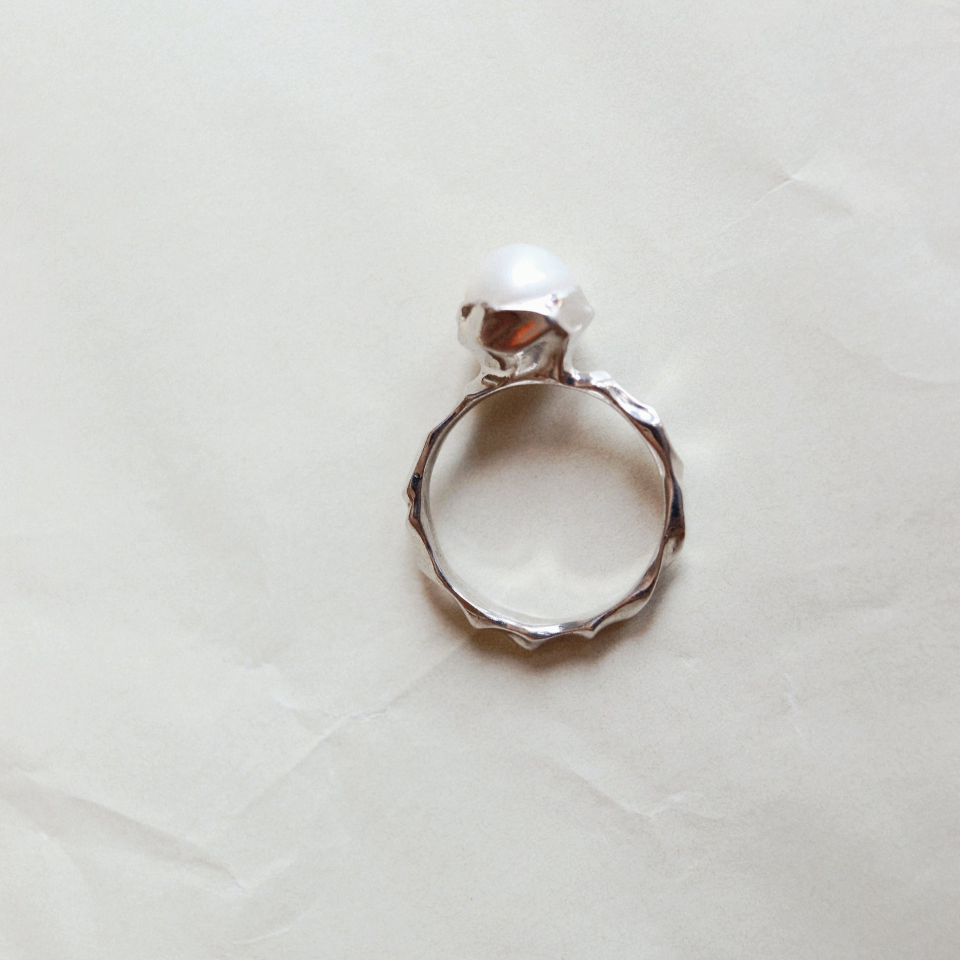 A silver ring with a textured, hammered band and a single white pearl set in a polished bezel, photographed on a wrinkled off-white paper background.