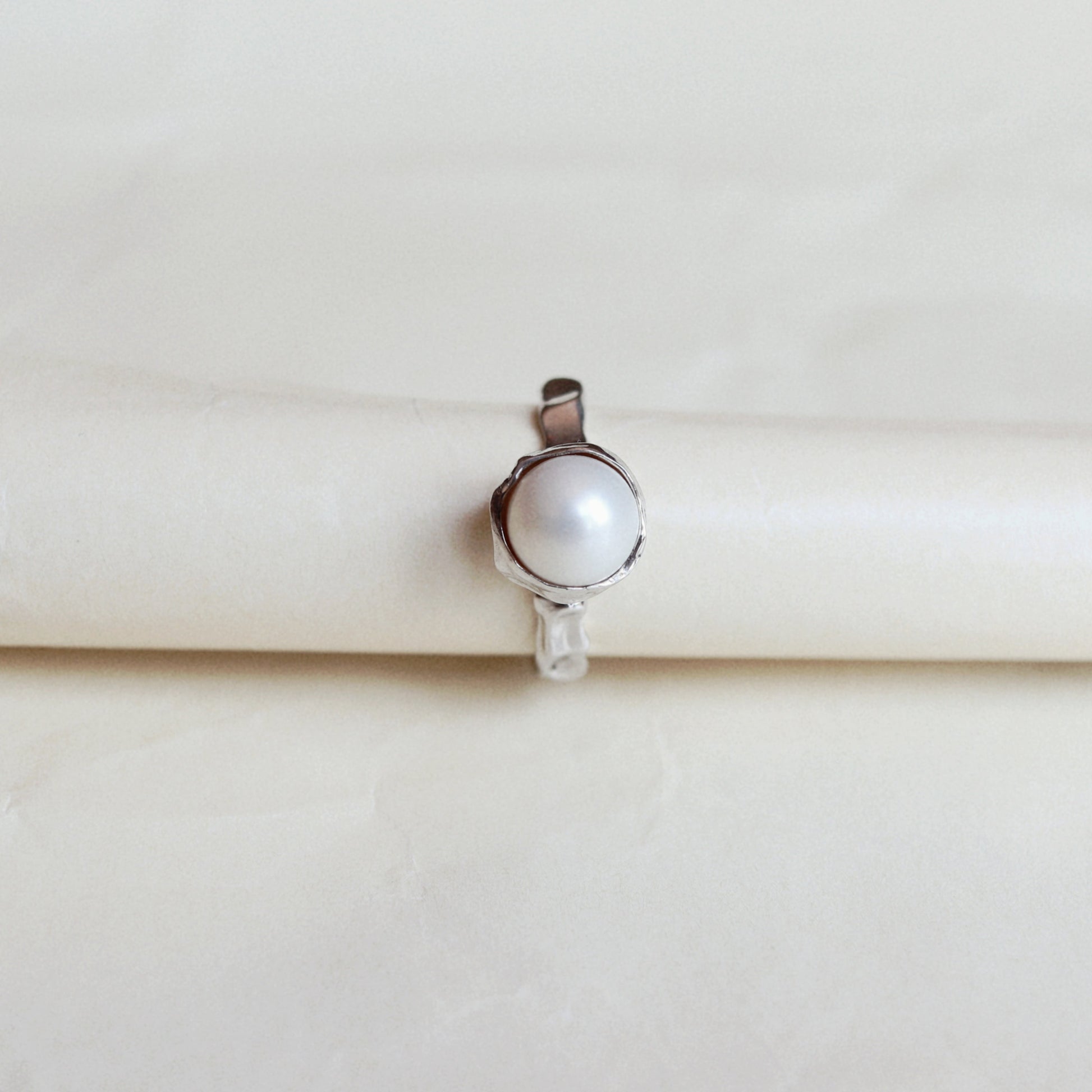 A silver ring featuring a single white pearl bezel-set in a polished silver band, displayed on a cream-colored ring holder.