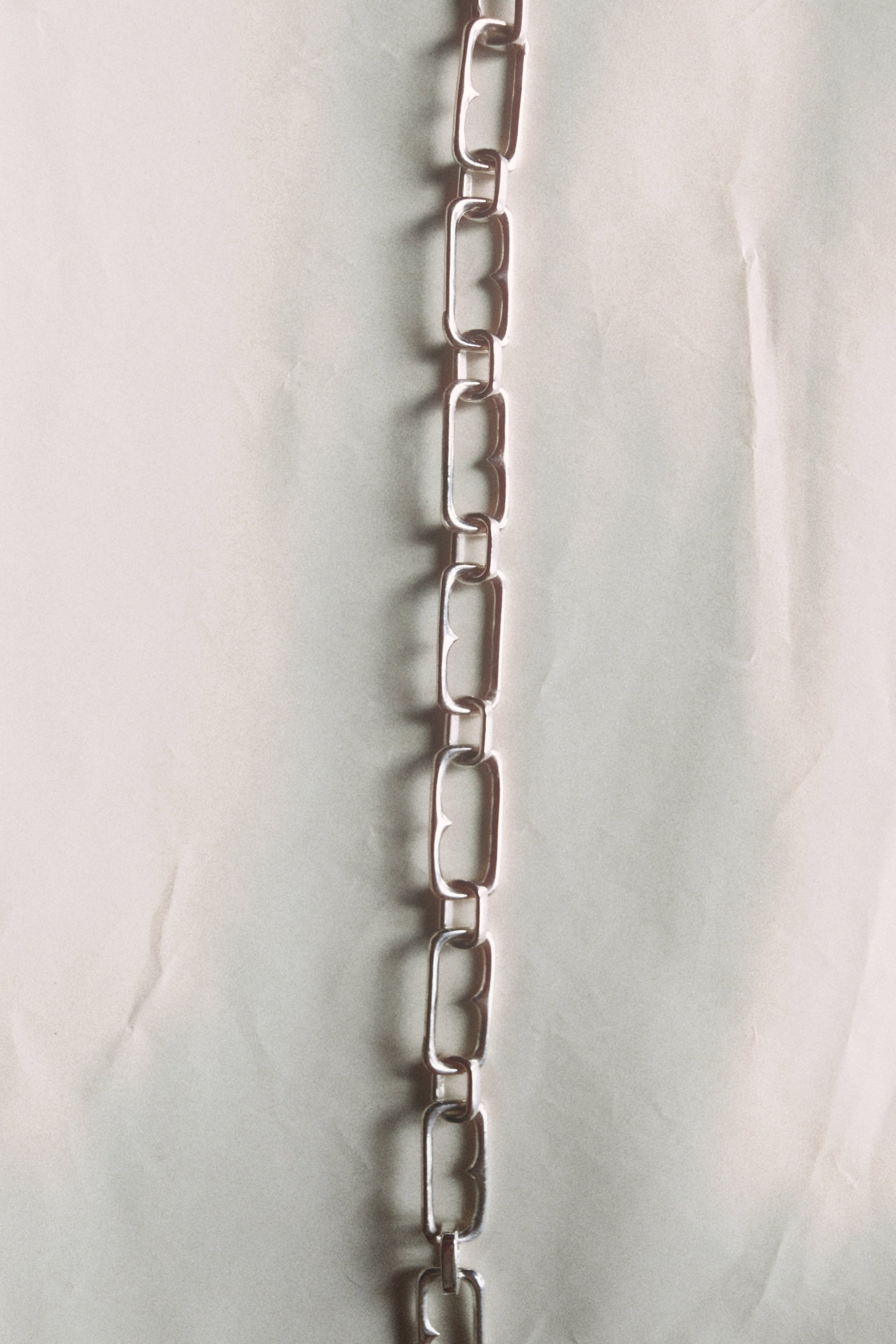 A vertical silver metal link chain with elongated rectangular links, photographed against a light off-white fabric background.