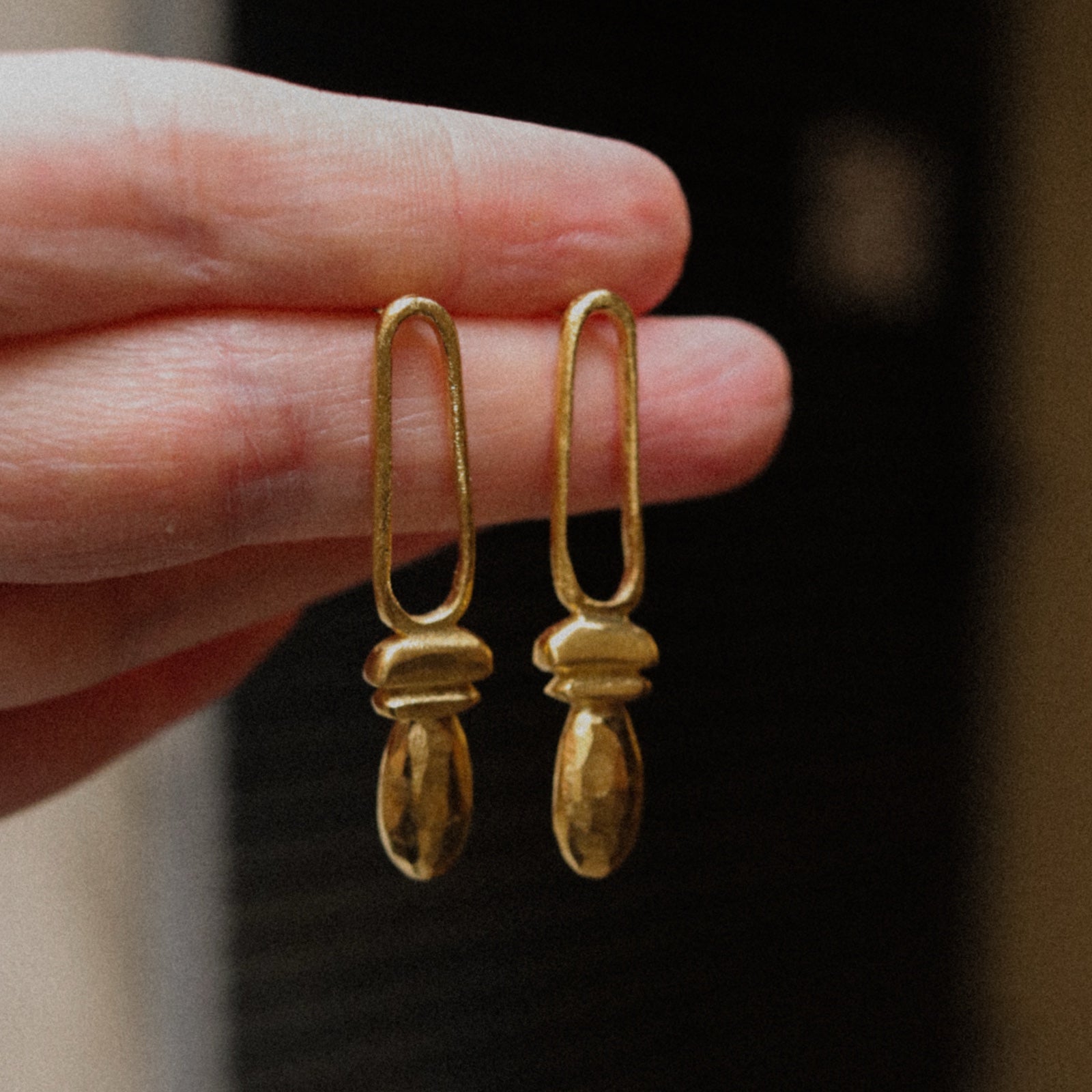 Gold earrings held between fingers against a dark background