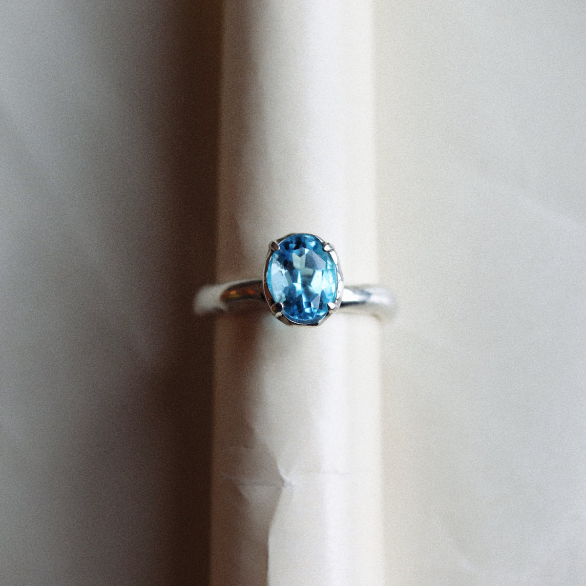 An oval-cut light blue gemstone ring set in a silver band, displayed on a cream-colored cylindrical stand against a neutral beige background.