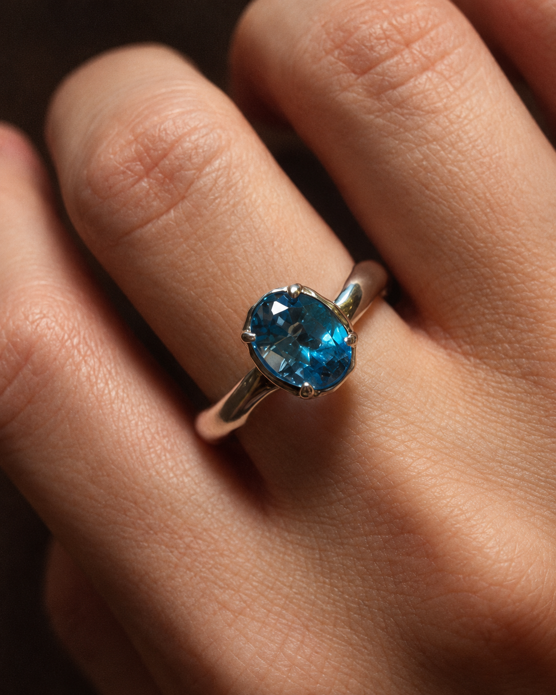 A close-up of a hand wearing a silver ring with a faceted oval blue gemstone.