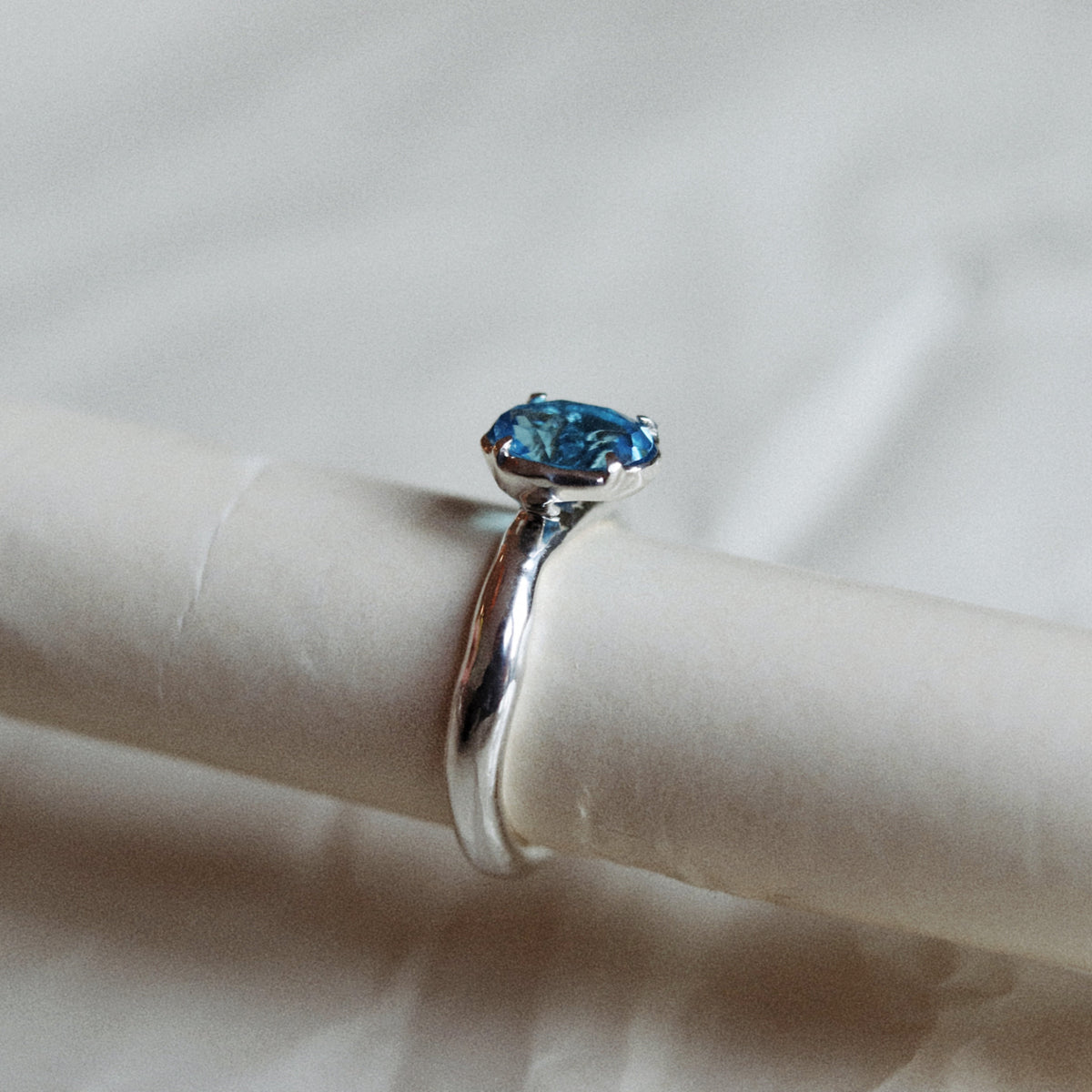 A silver ring with a marquise-cut blue gemstone displayed on a white ring stand against a soft, light gray fabric background.