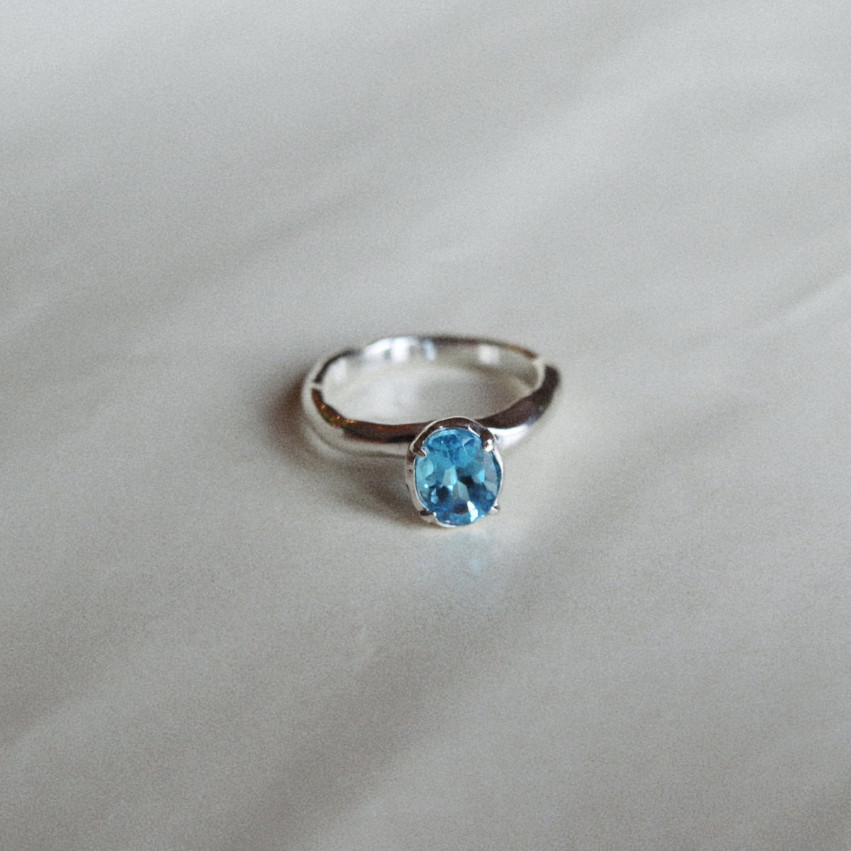 A silver ring with a small oval-cut light blue gemstone, resting on a soft, light gray fabric background.