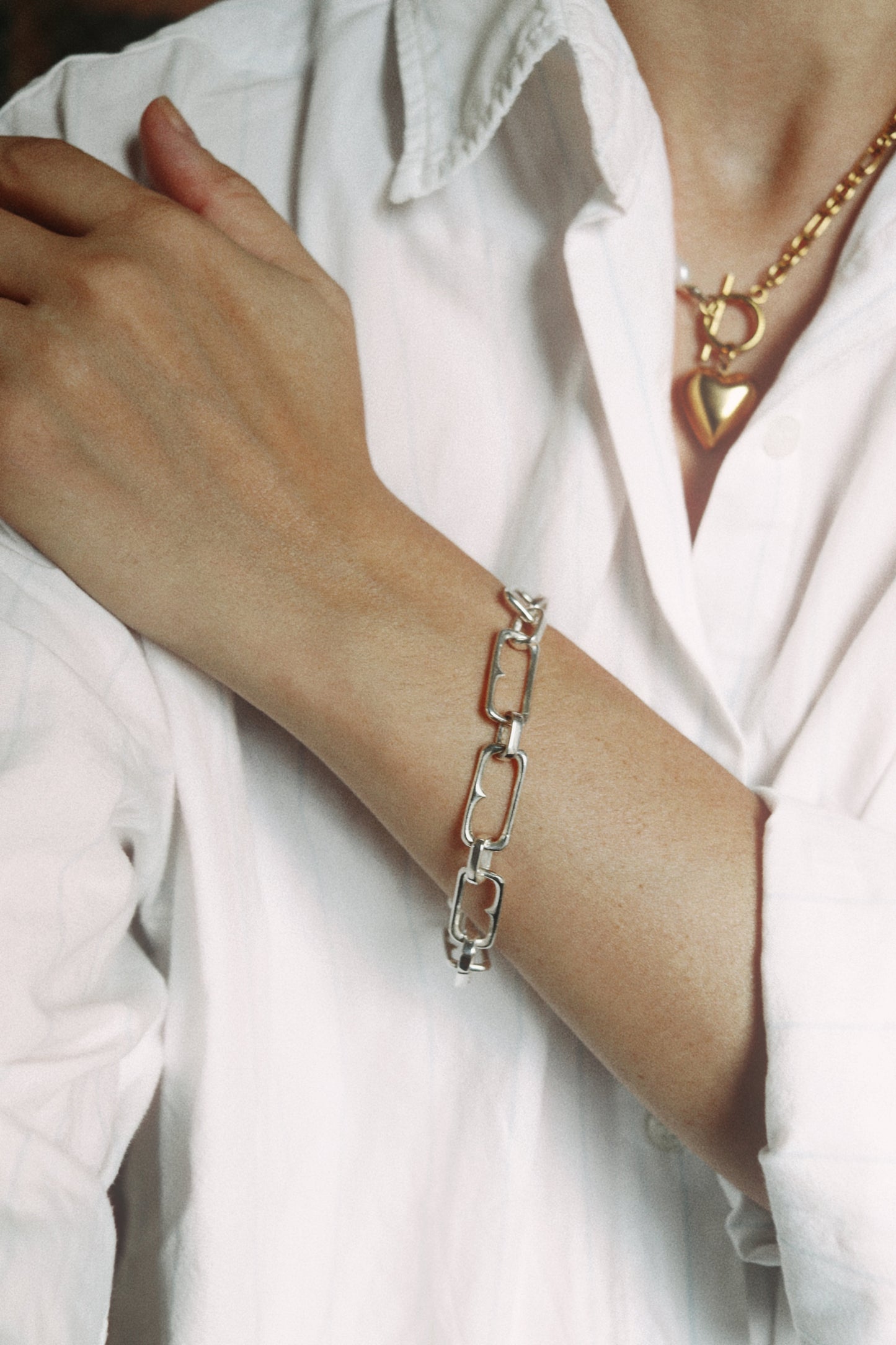 A silver chain link bracelet worn on a person's wrist, paired with a gold heart pendant necklace and a white button-up shirt.