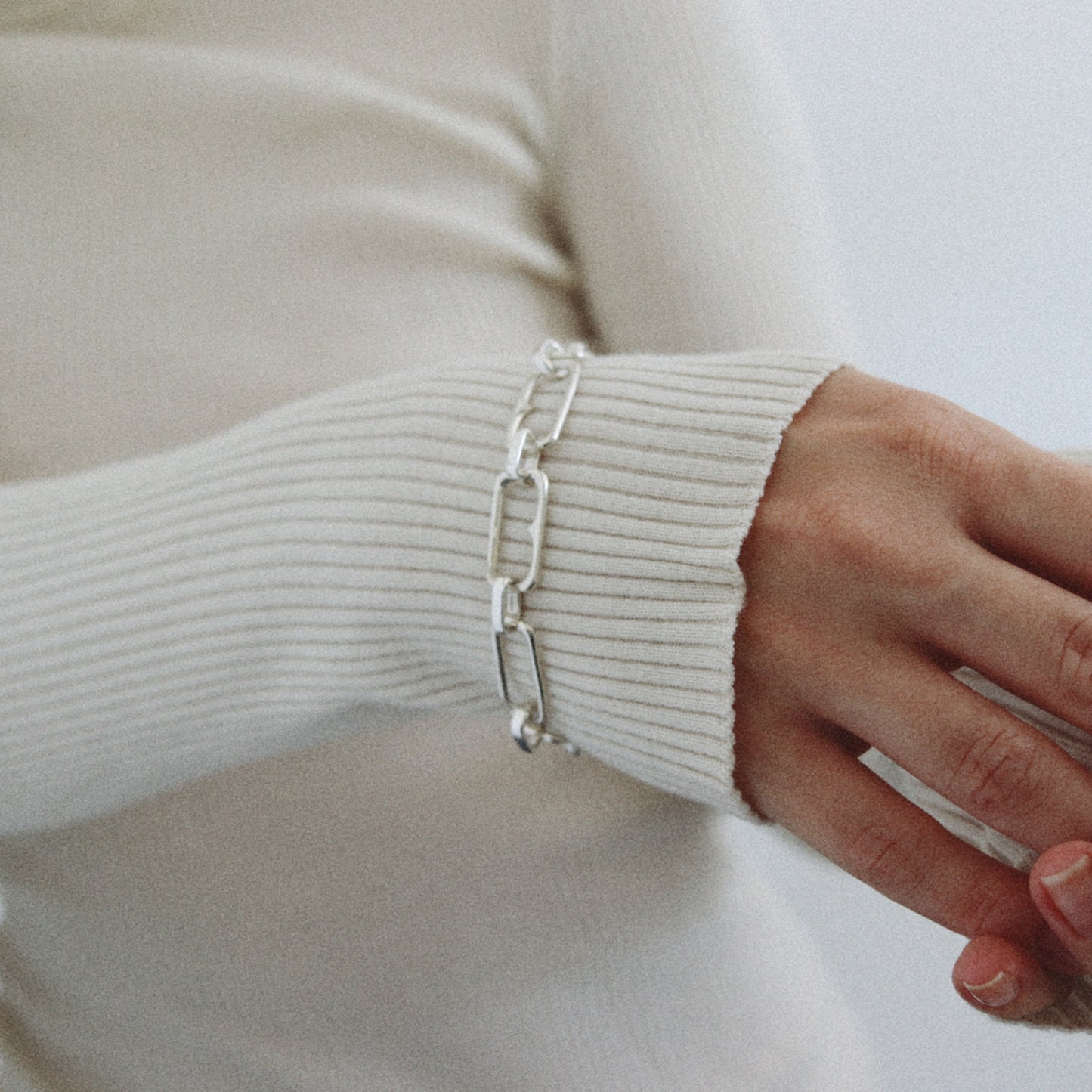 A close-up of a person wearing a silver chain link bracelet on their wrist, dressed in a cream ribbed knit sweater against a soft light gray background.