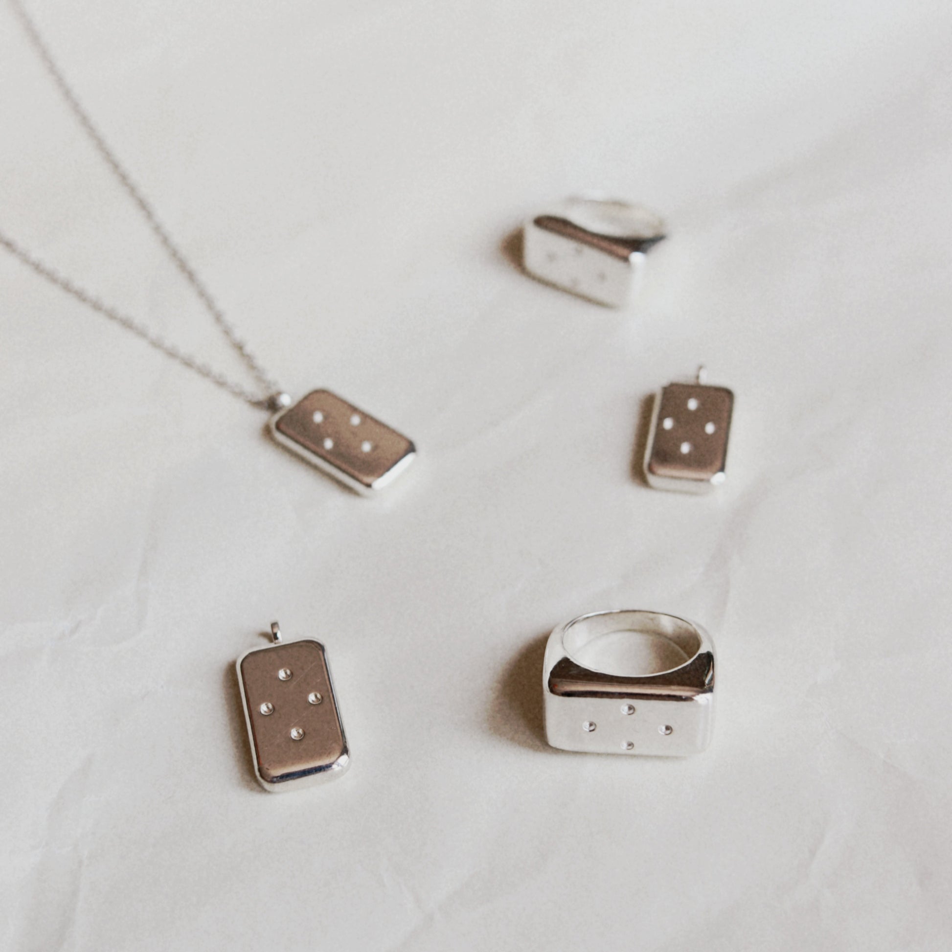 A set of minimalist silver jewelry pieces featuring rectangular pendants and rings with small white dot patterns, displayed on a textured white fabric background.