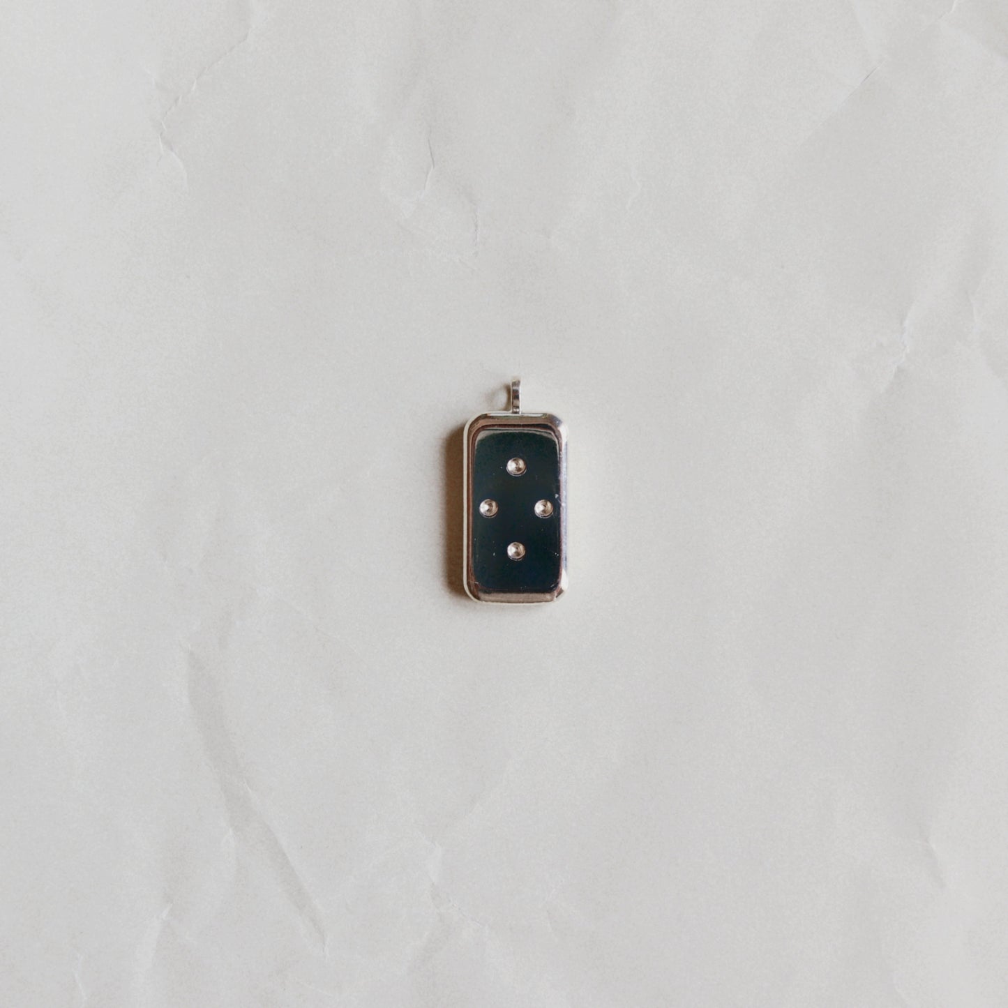 A small rectangular pendant with a dark navy blue enamel face and four silver-tone dots, set in a silver-tone metal frame, displayed on a textured white paper background.