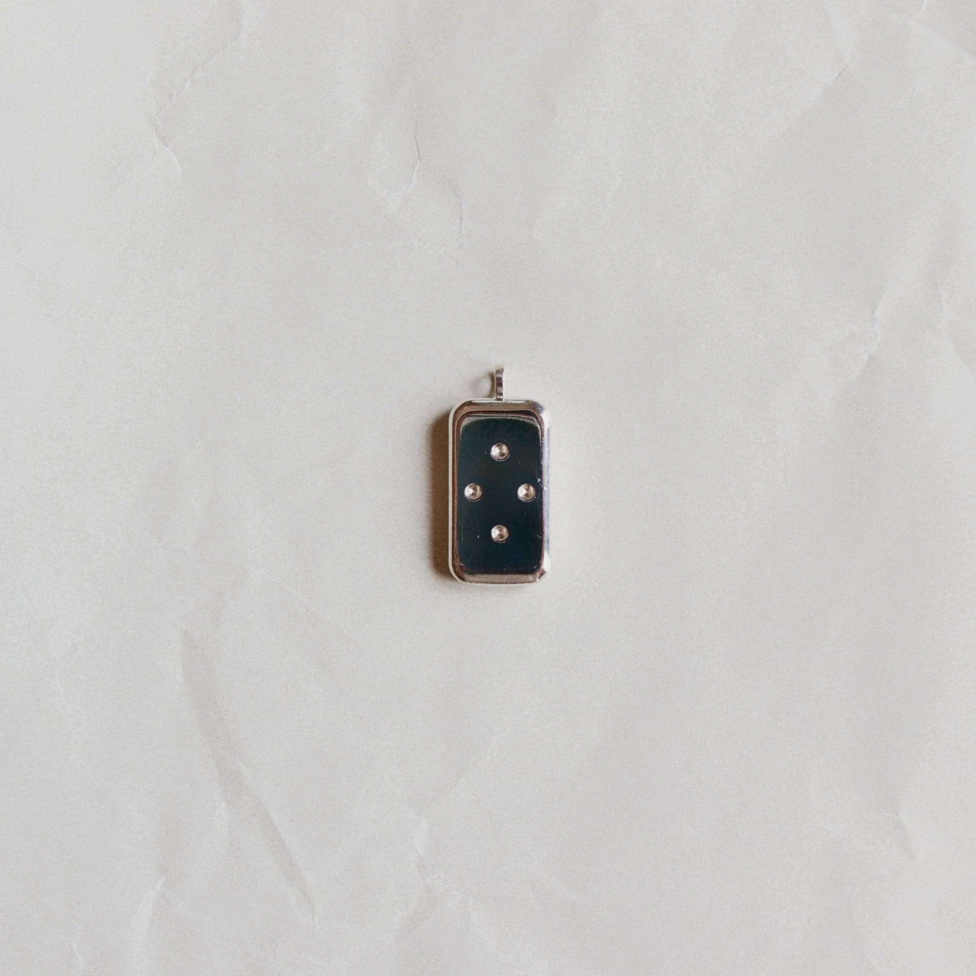 A small rectangular pendant with a dark navy blue enamel face and four silver-tone dots, set in a silver-tone metal frame, displayed on a textured white paper background.