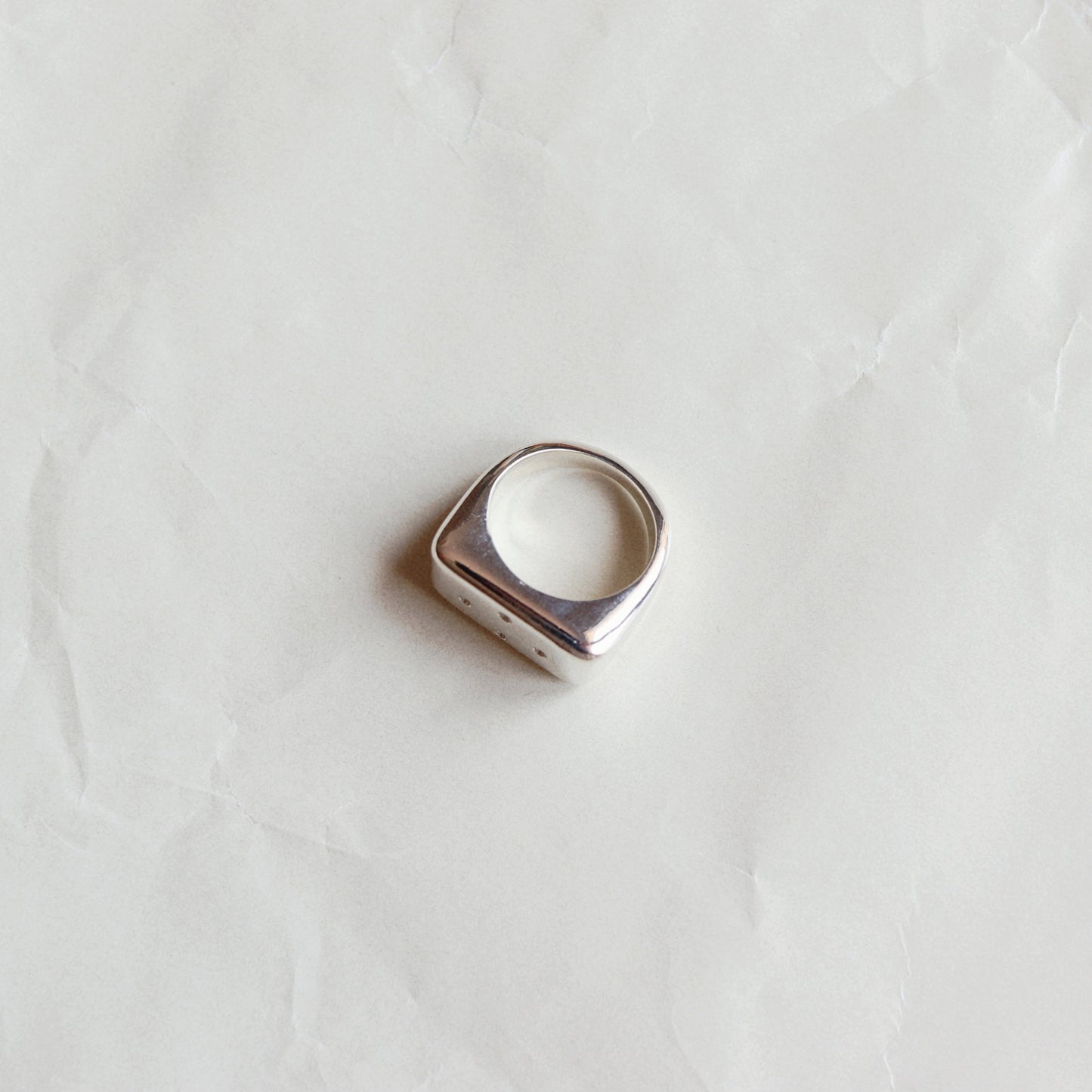A silver-toned signet ring with a wide, flat rectangular face, photographed on a textured, off-white paper background.