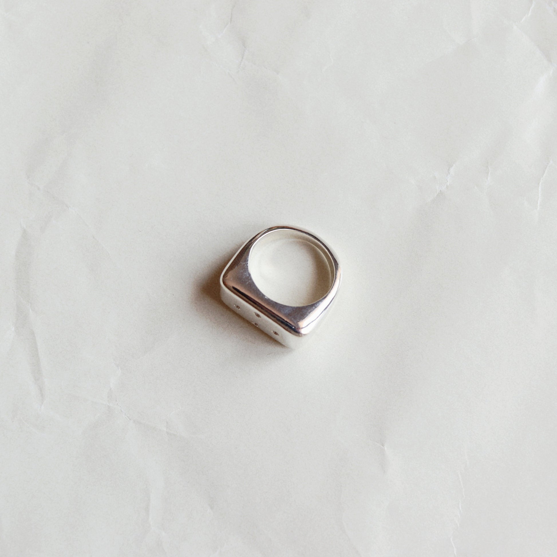 A silver-toned signet ring with a wide, flat rectangular face, photographed on a textured, off-white paper background.