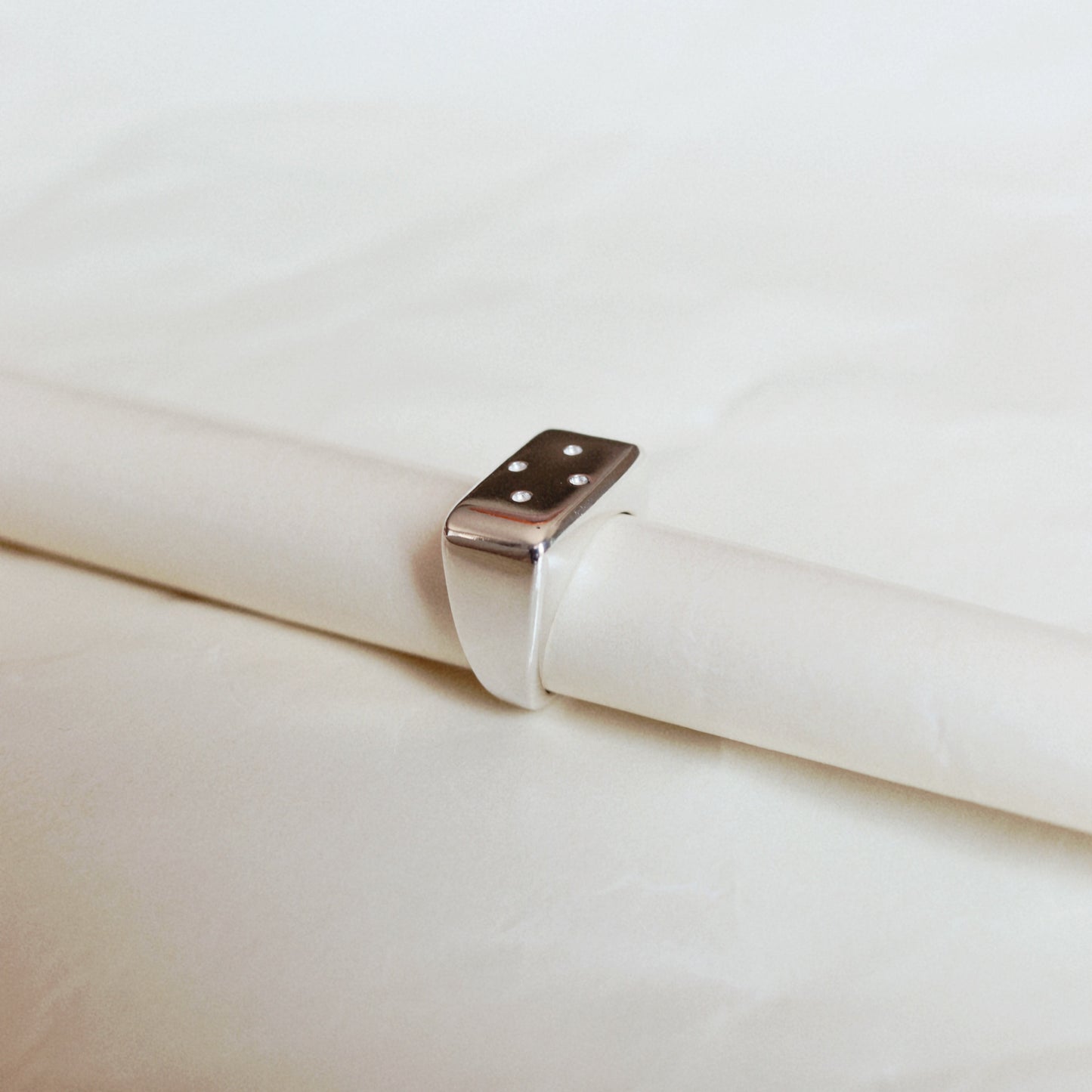 A silver ring with a rectangular, rounded-corner top featuring three small dots, shown resting on a white cylindrical display against a soft white background.