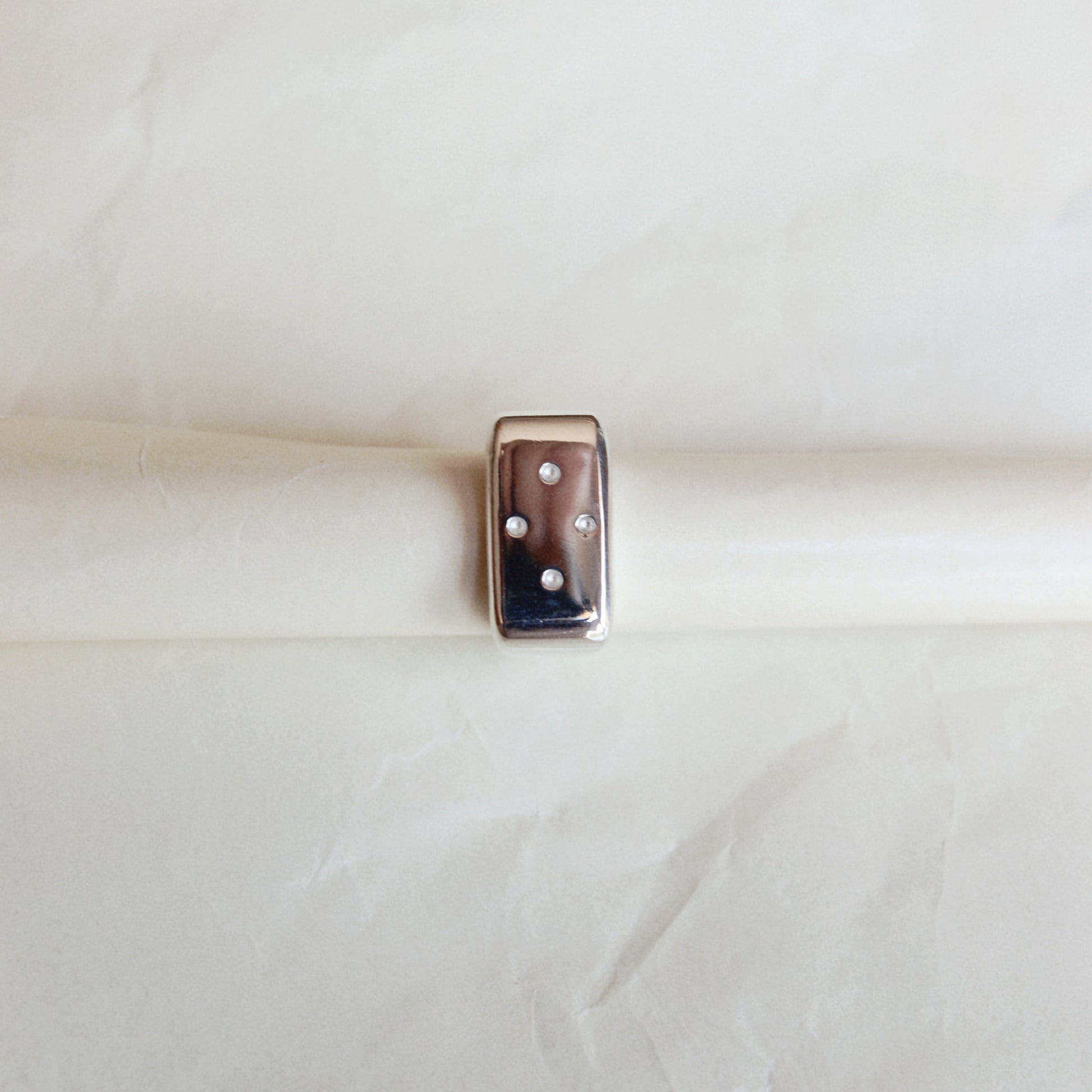 A small rectangular metallic charm with a glossy finish and three white dots, resting horizontally on a textured off-white paper background.