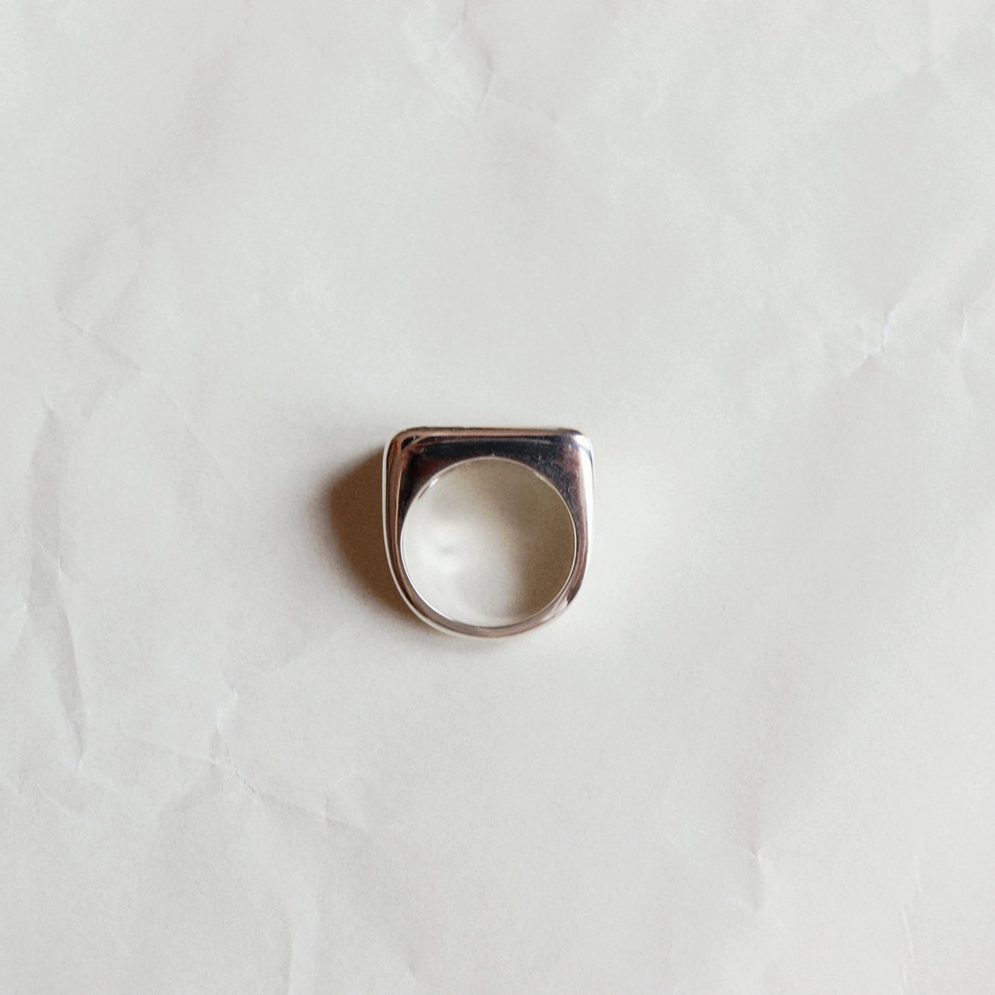 A silver-toned signet ring with a square face and rounded corners, displayed on a textured, off-white paper background.