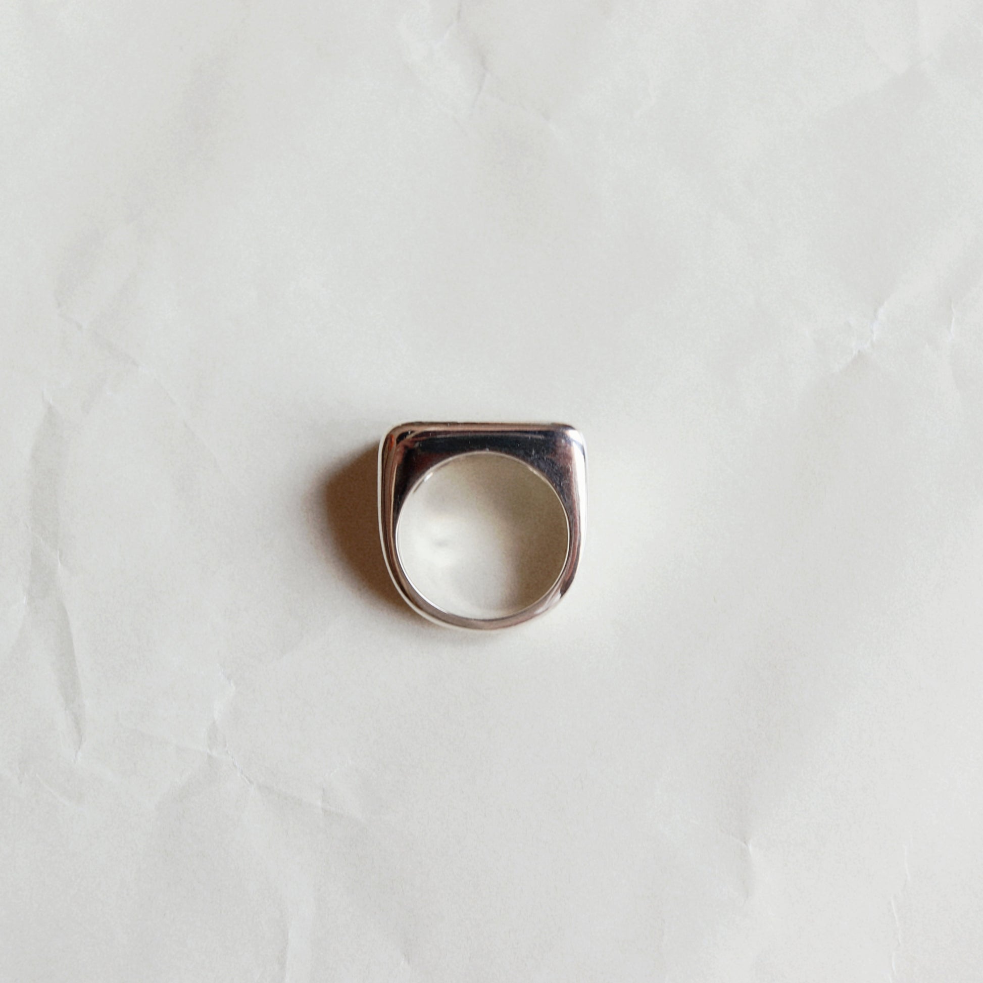 A silver-toned signet ring with a square face and rounded corners, displayed on a textured, off-white paper background.