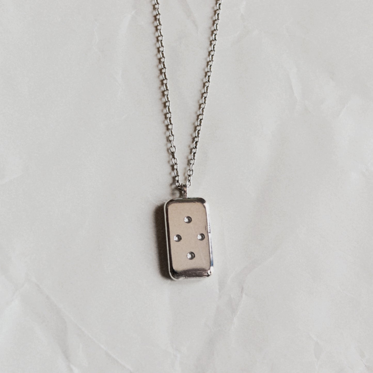 A silver-toned rectangular pendant necklace with four small dots, suspended from a matching chain against a wrinkled off-white background.