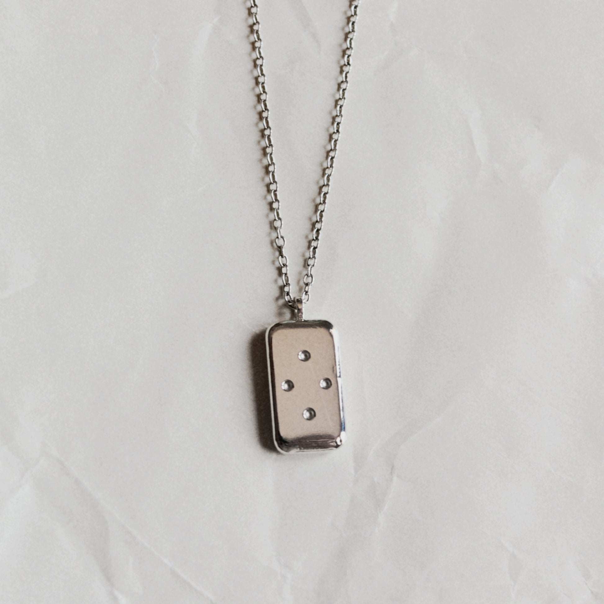 A silver-toned rectangular pendant necklace with four small dots, suspended from a matching chain against a wrinkled off-white background.