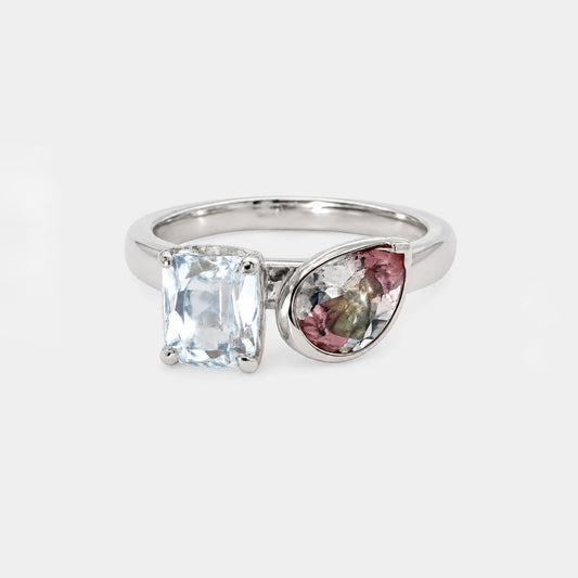 A silver ring featuring a rectangular clear gemstone and a teardrop-shaped multicolor gemstone cluster, set against a light gray background.
