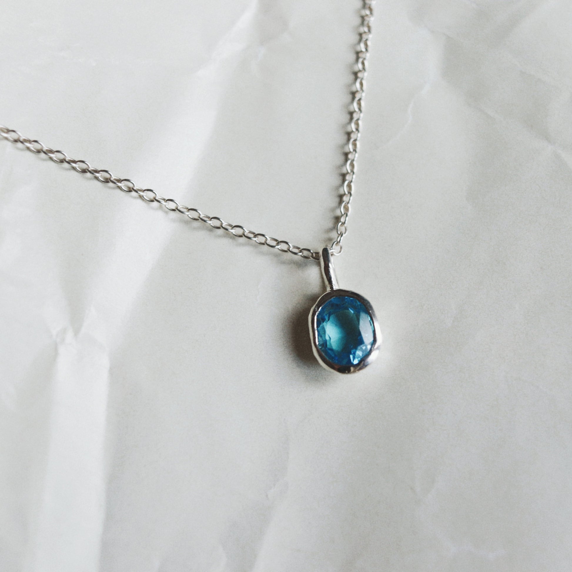 A silver chain necklace with a small oval blue gemstone pendant, displayed on a crumpled white paper background.