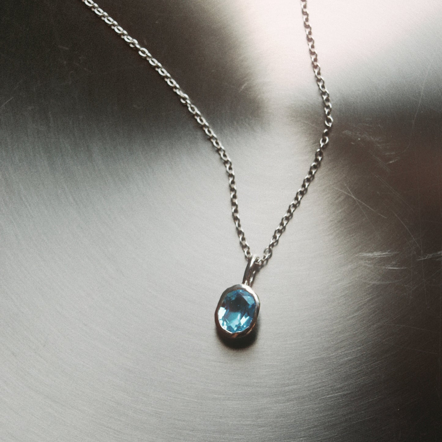 A silver chain necklace with a small oval-cut light blue gemstone pendant, displayed on a brushed metallic surface.