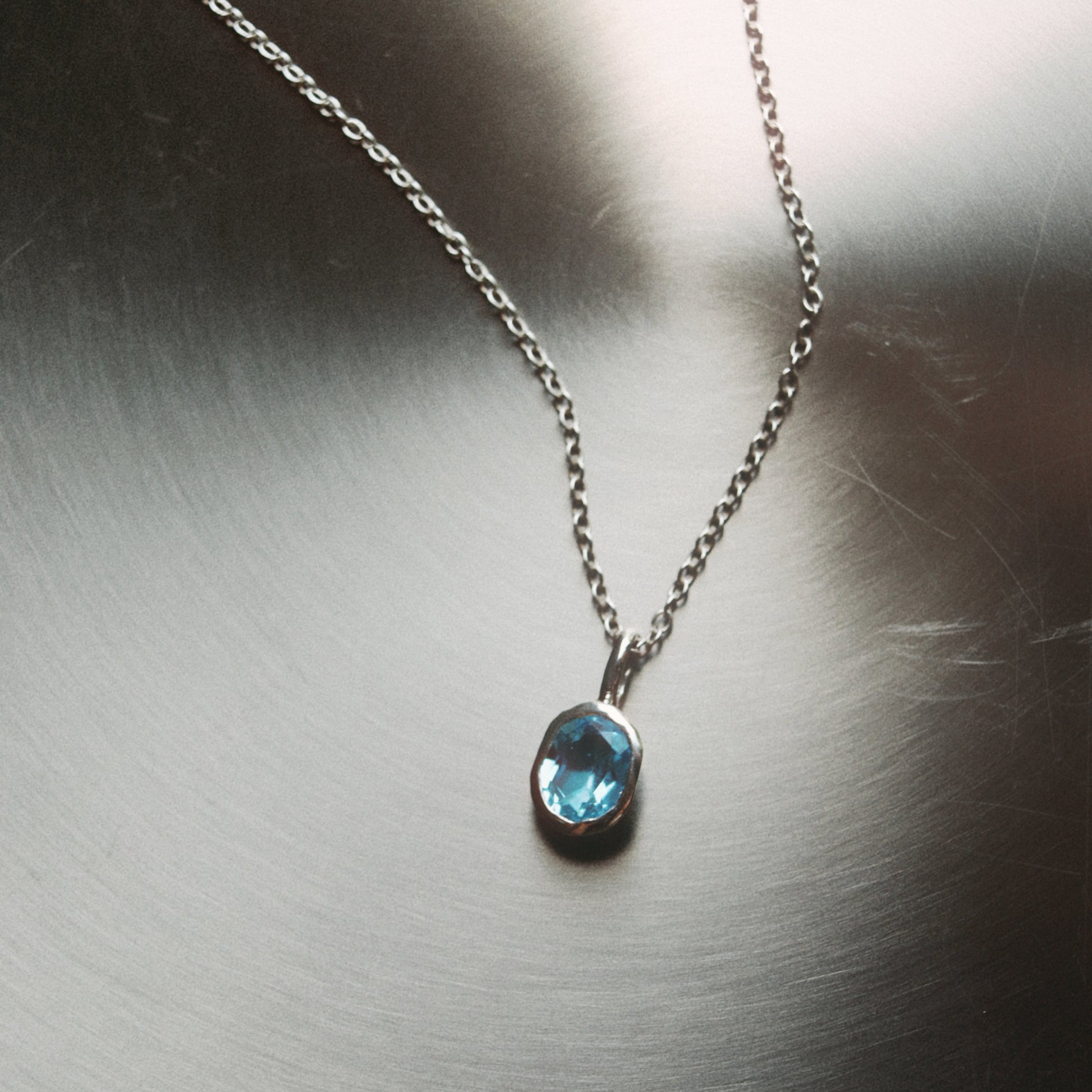 A silver chain necklace with a small oval-cut light blue gemstone pendant, displayed on a brushed metallic surface.