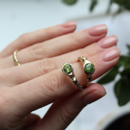 Close-up of a hand wearing two gold rings with green gemstones against a blurred background.