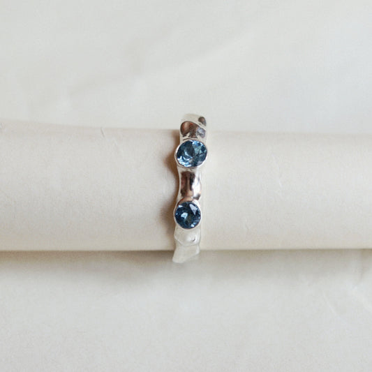 A silver ring with two round blue gemstones, displayed on a cream-colored ring stand against a light background.