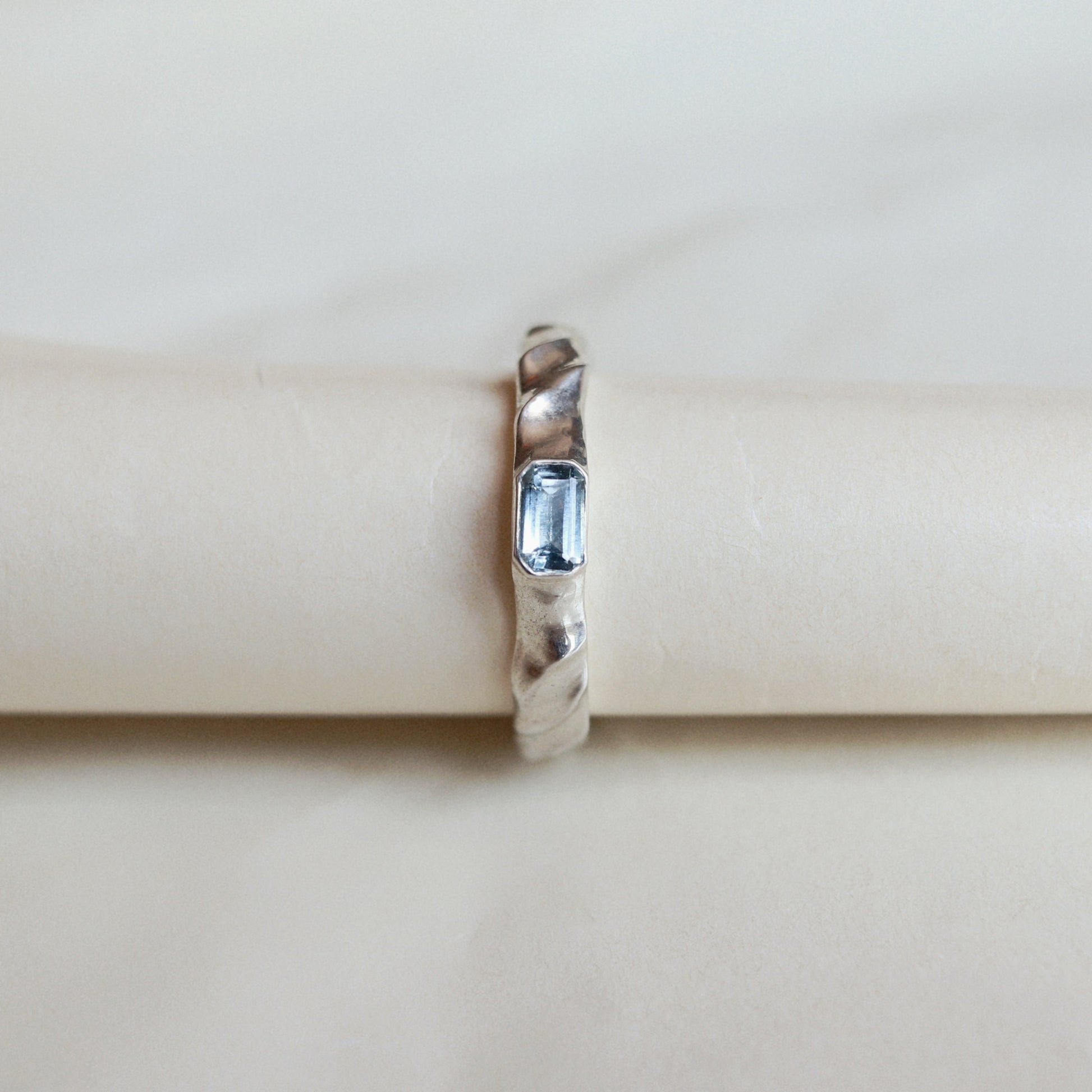 A silver ring with a textured twisted band and a rectangular blue gemstone, displayed on a cream-colored ring stand.