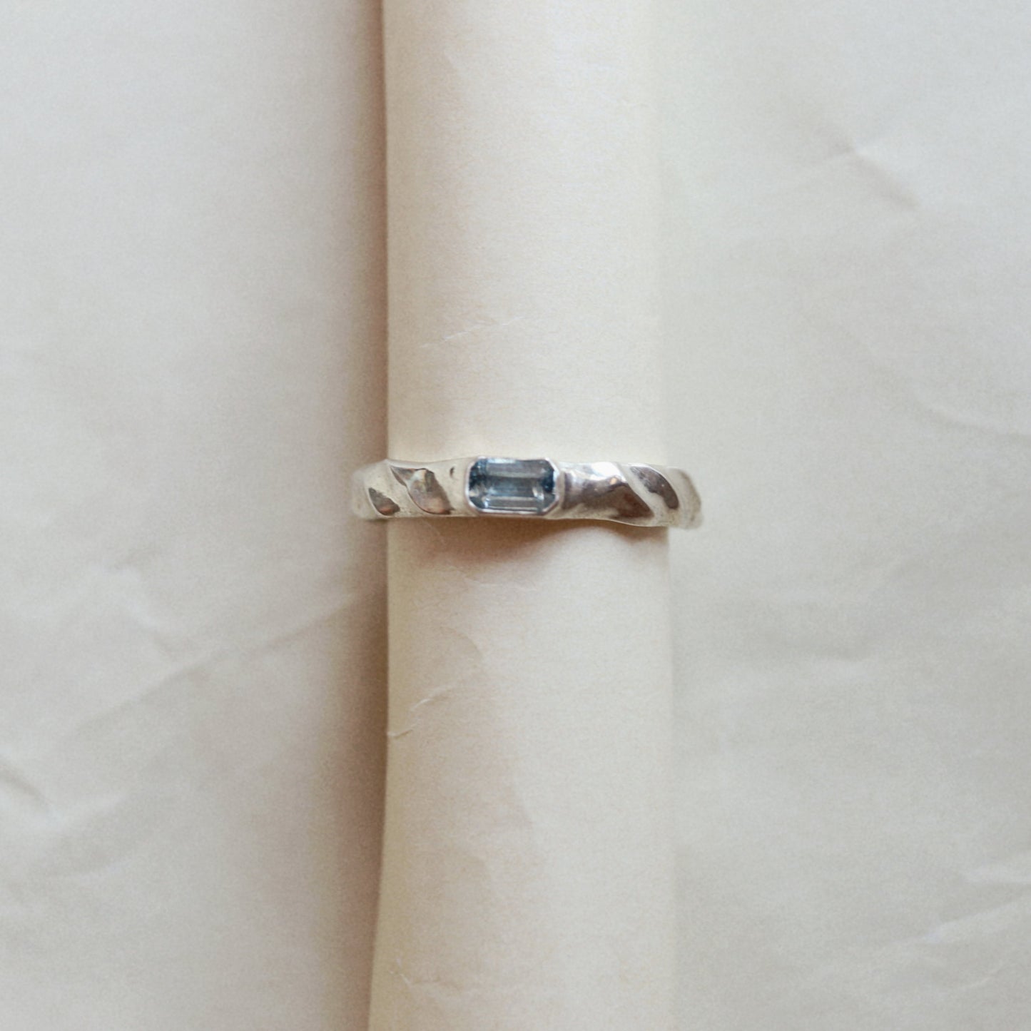 A silver ring with a rectangular blue gemstone, displayed on a rolled cream-colored paper band against a neutral light background.