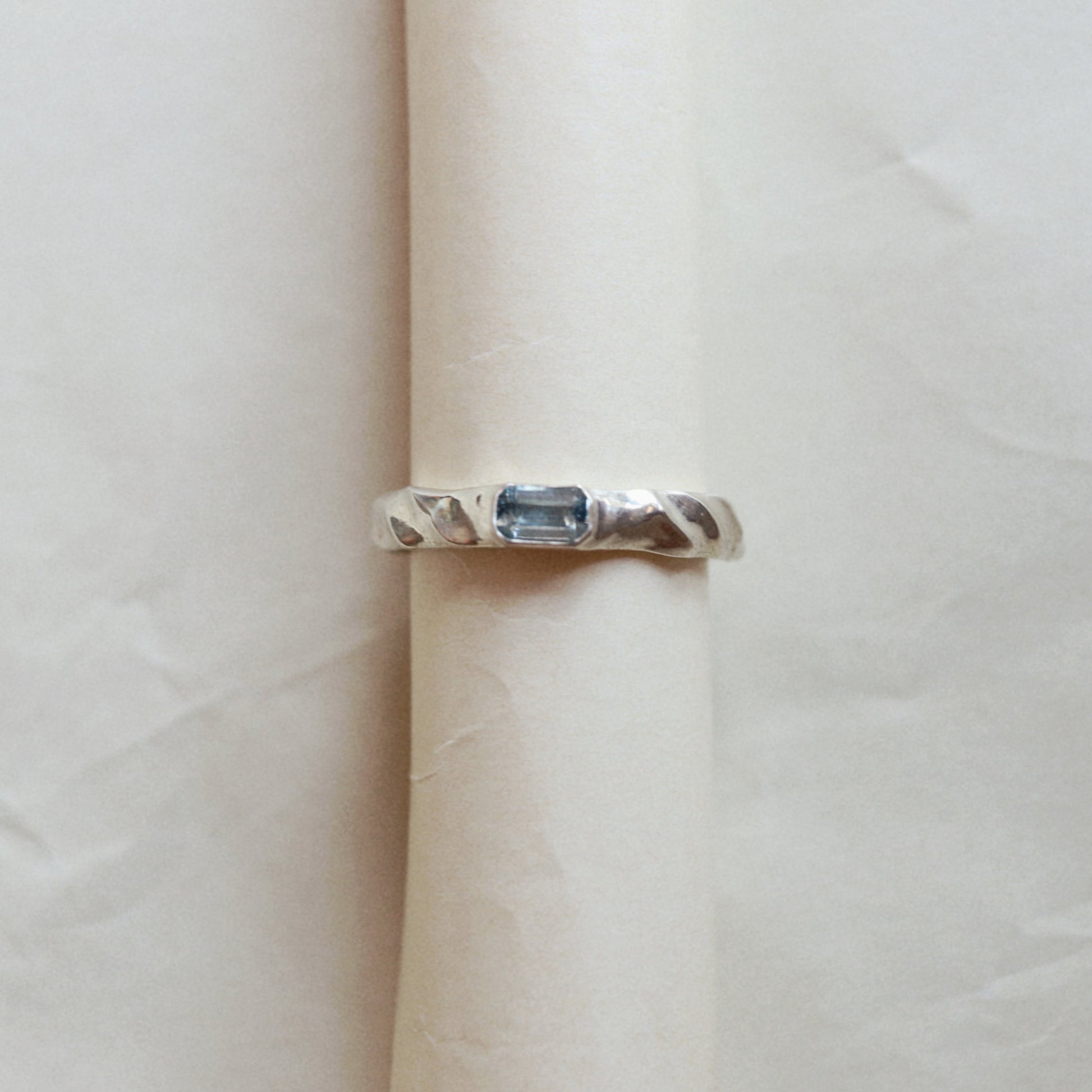 A silver ring with a rectangular blue gemstone, displayed on a rolled cream-colored paper band against a neutral light background.
