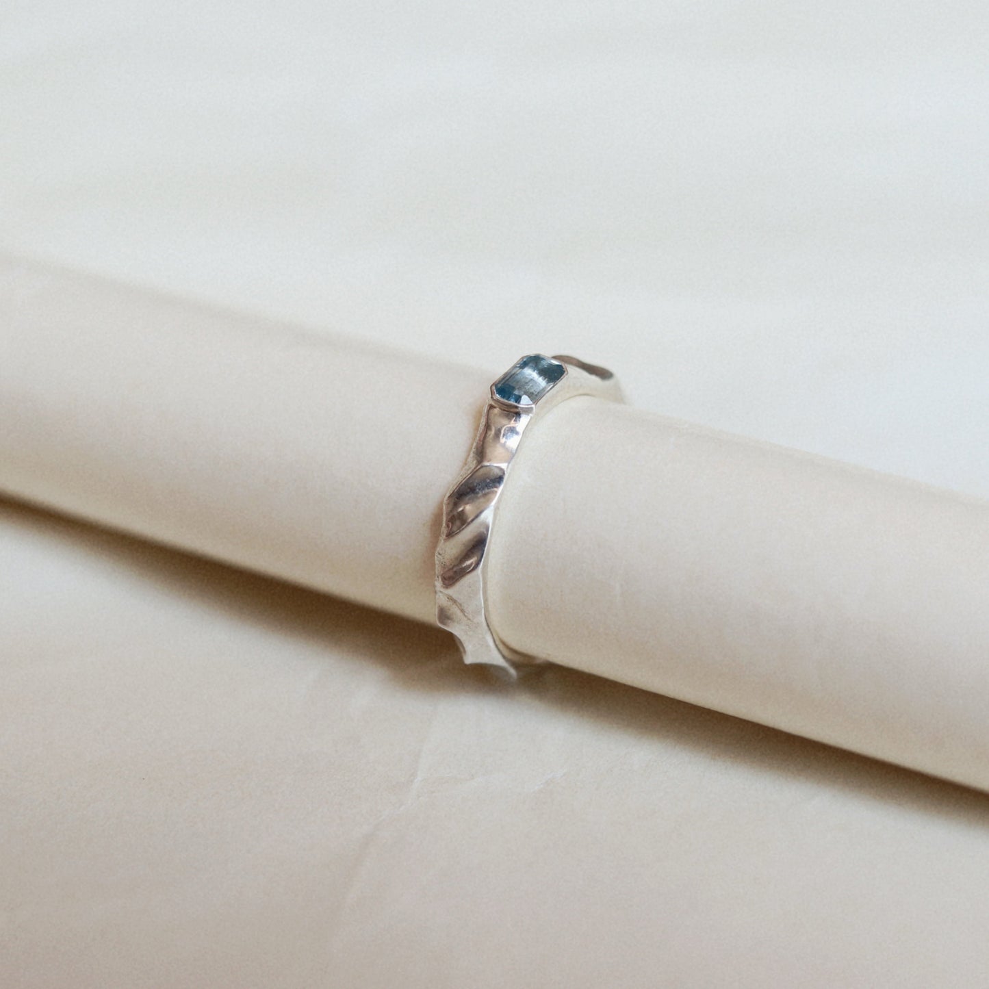 A silver twisted band ring with a small rectangular blue gemstone, displayed on a cream-colored paper roll against a light beige background.
