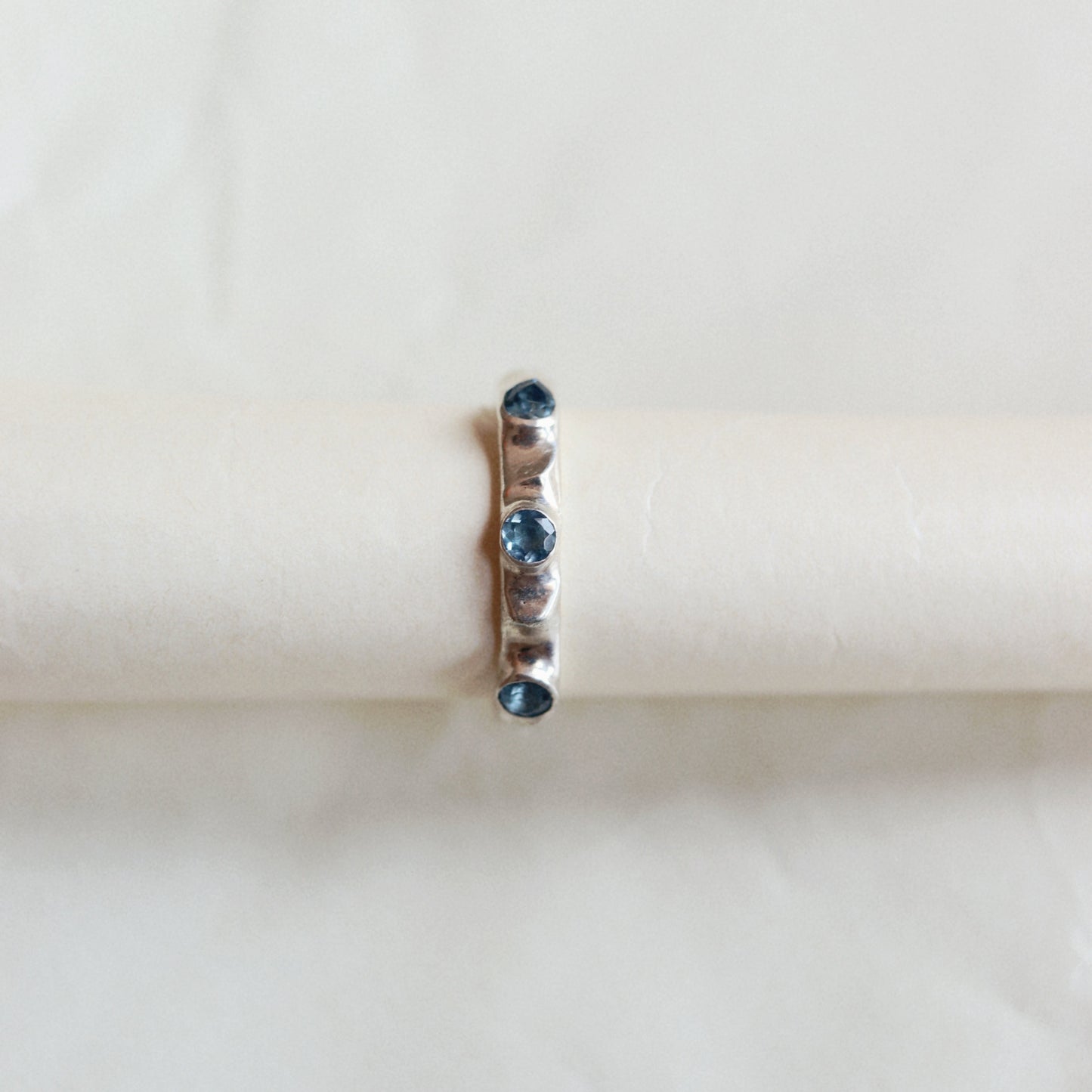 A delicate ring with small blue gemstones and a central clear stone, displayed on a cream-colored ring stand against a soft, light background.