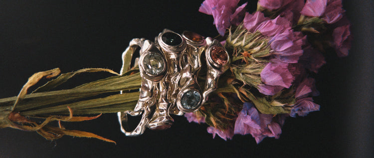 Silver ring with gemstones held by dried flowers against a black background