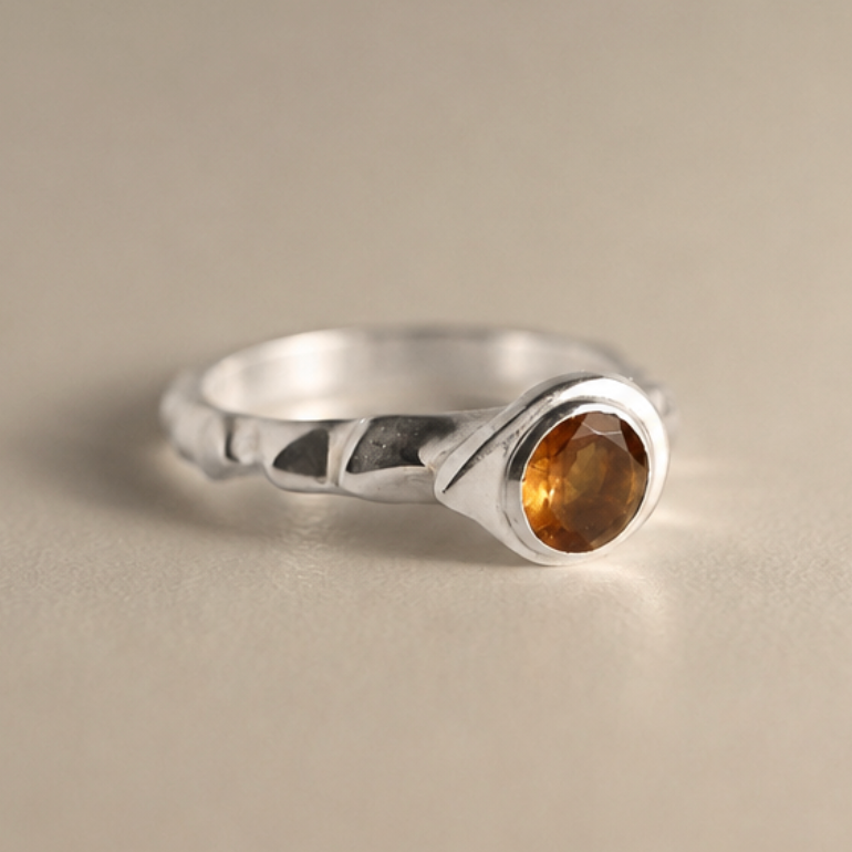 A sterling silver ring with a textured band and an oval-cut amber gemstone, photographed on a neutral beige background.