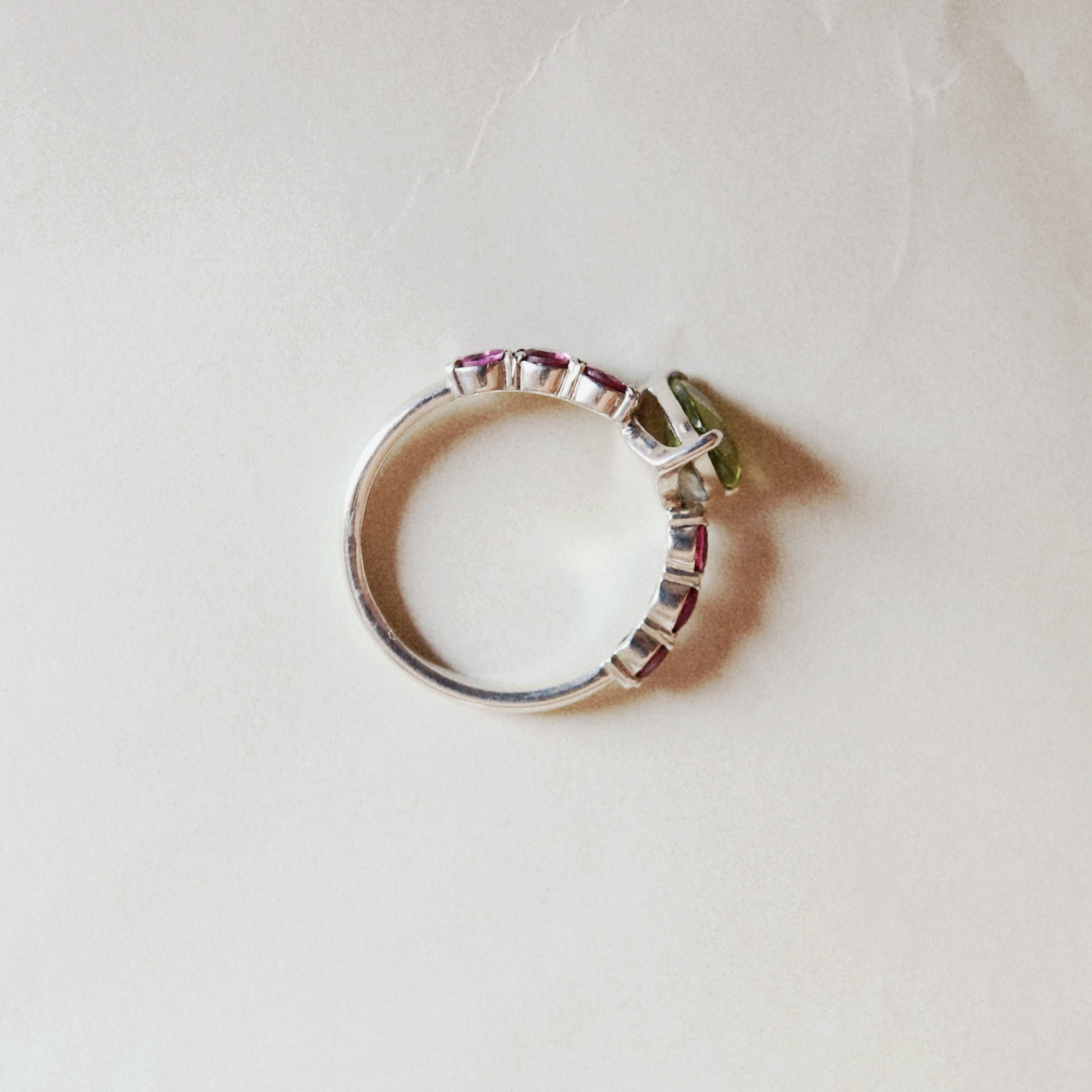 A silver band ring featuring a small green gemstone and a row of pink stones, photographed on a textured off-white surface.