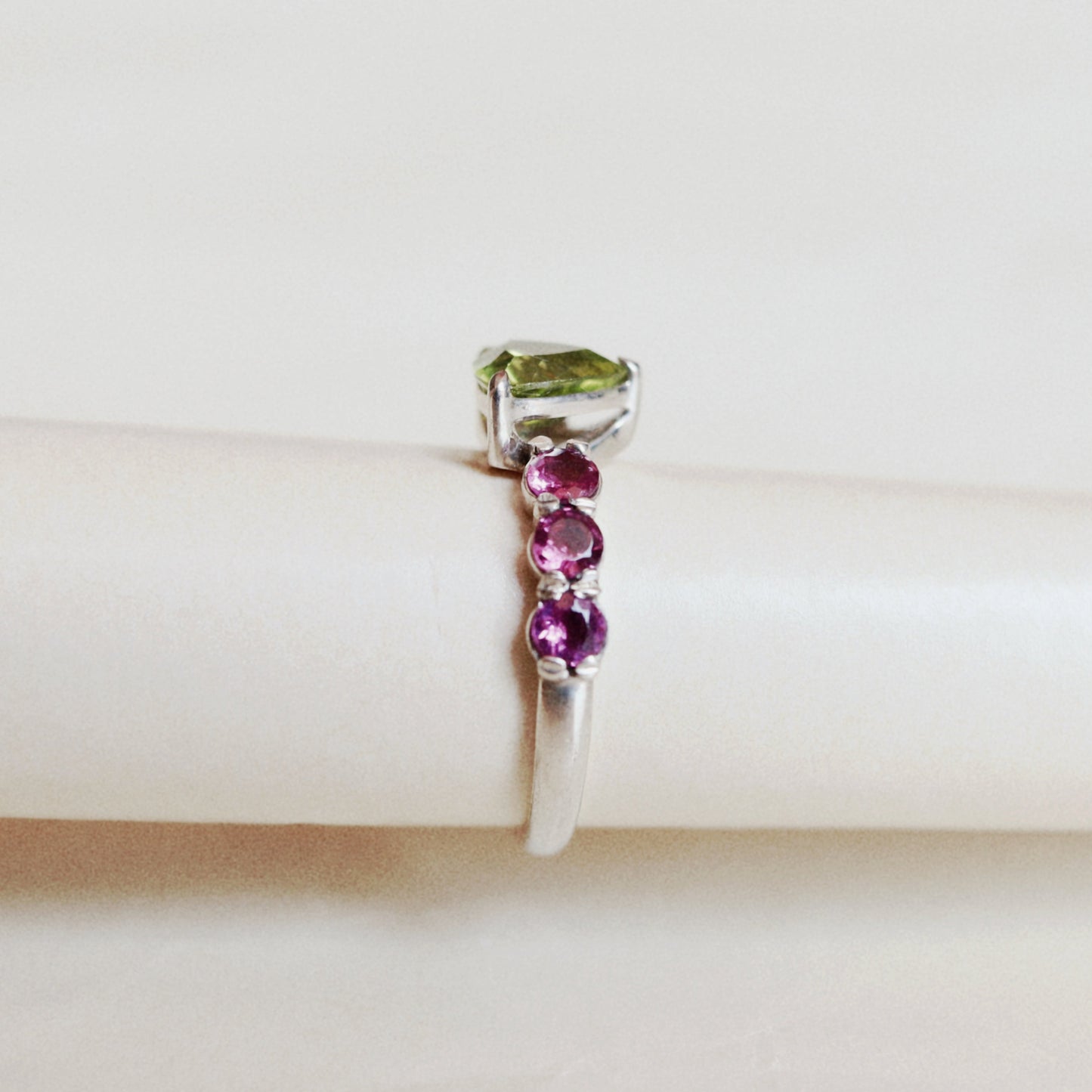A silver ring featuring a rectangular green gemstone at the top and three round pink gemstones set vertically along the band, displayed on a cream-colored ring stand.