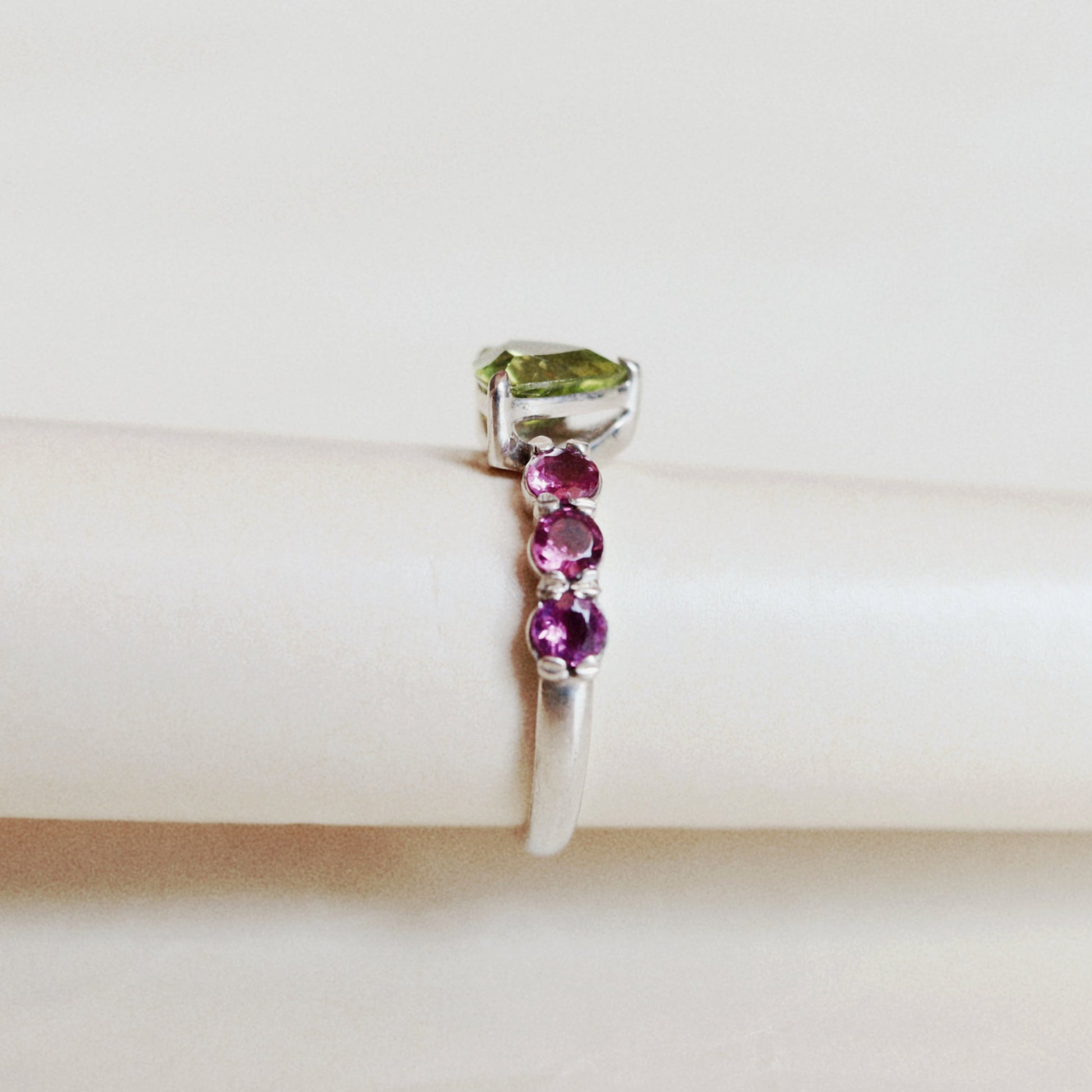 A silver ring featuring a rectangular green gemstone at the top and three round pink gemstones set vertically along the band, displayed on a cream-colored ring stand.