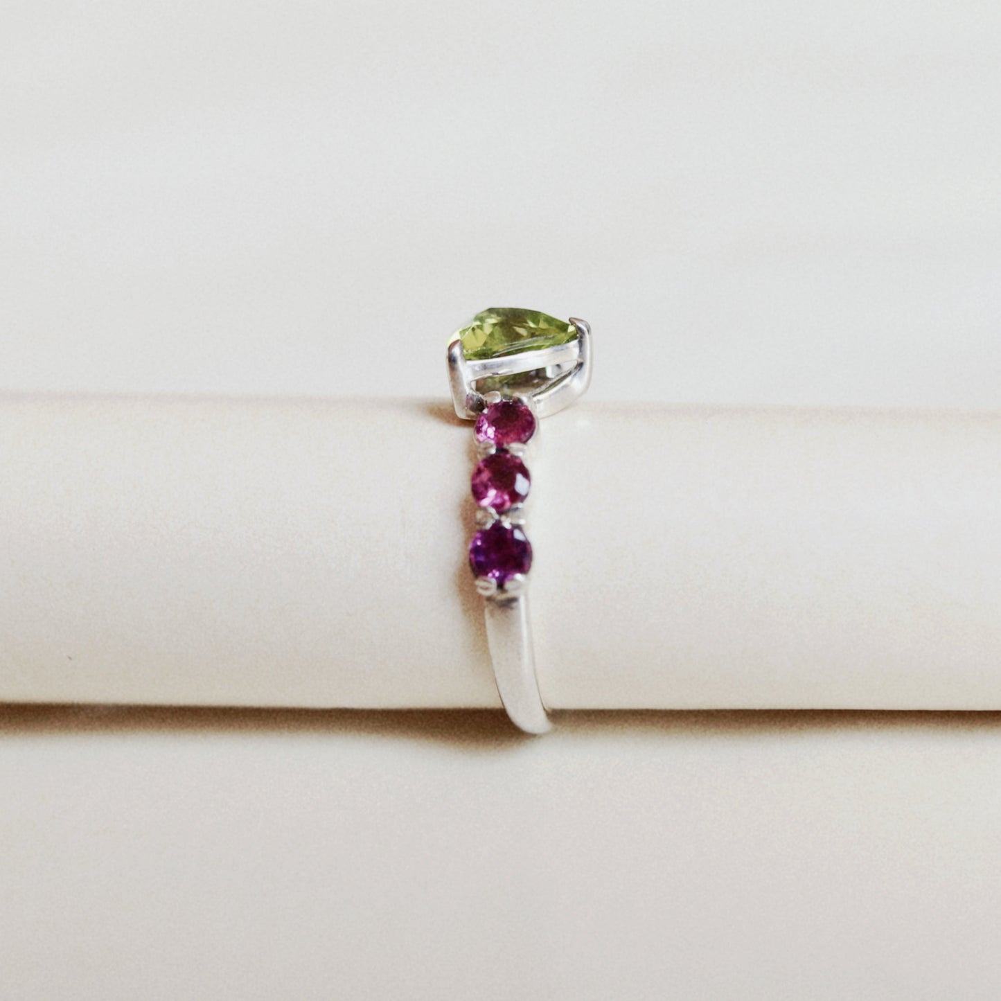 A silver ring displayed on a cream-colored ring holder. The ring features a triangular light green gemstone at the top and a vertical row of three round magenta-pink gemstones on a white band.