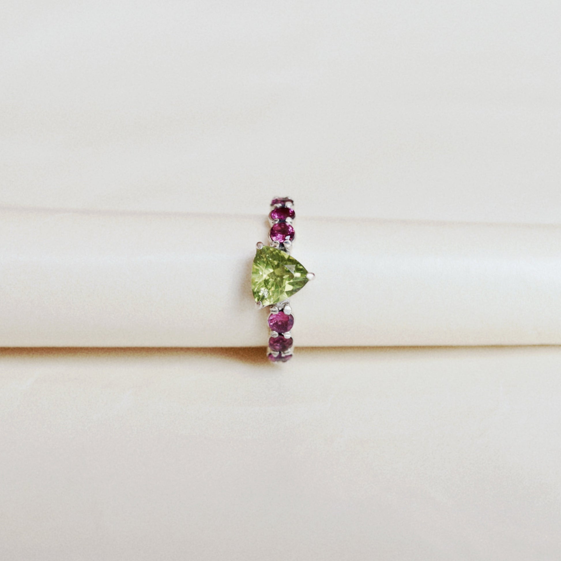 A delicate silver ring featuring a central green heart-shaped gemstone flanked by small pink gemstones on either side, displayed on a white cylindrical stand against a light beige background.