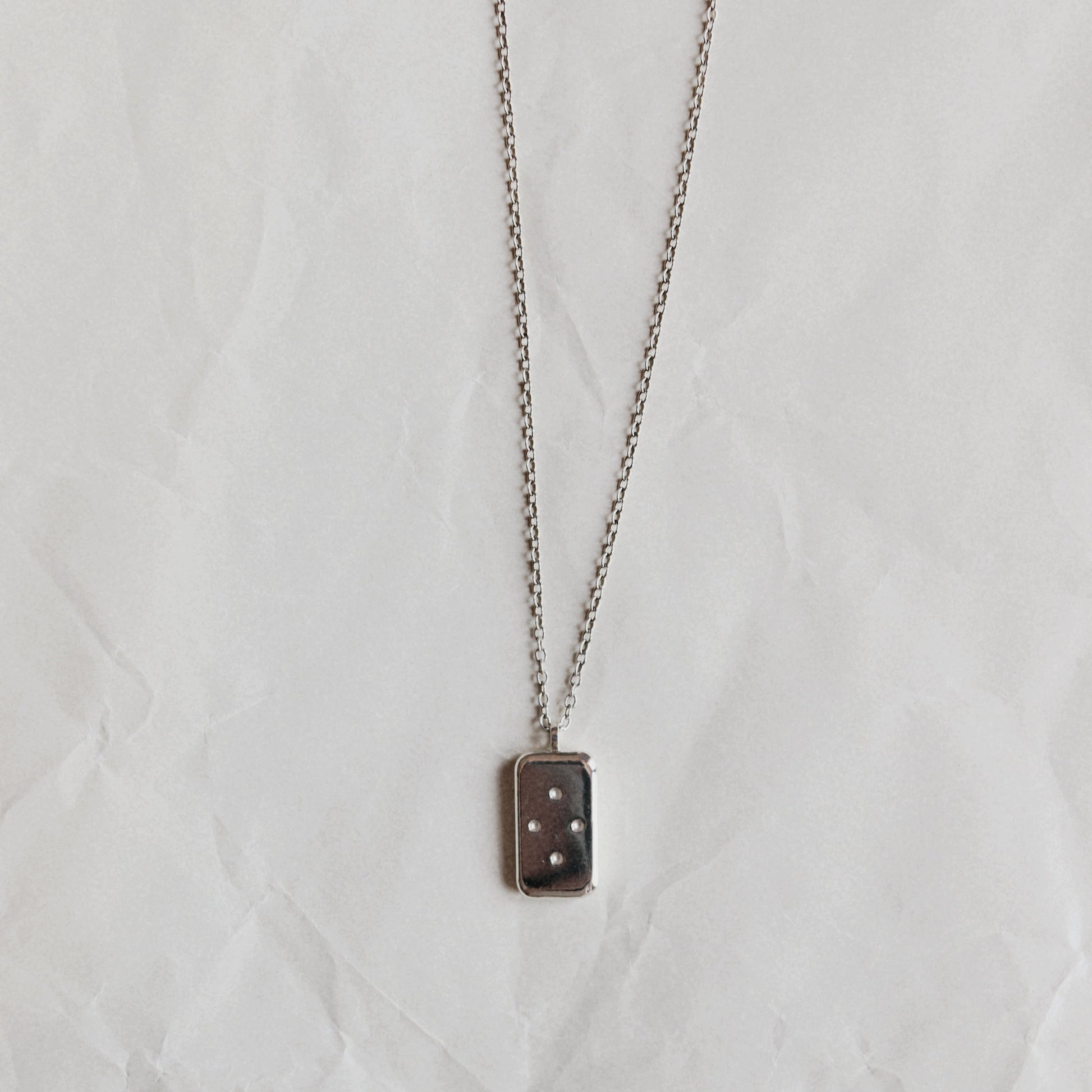 A silver-toned chain necklace with a small rectangular pendant featuring a dark brown or black background and three white dots, displayed on a wrinkled white paper background.